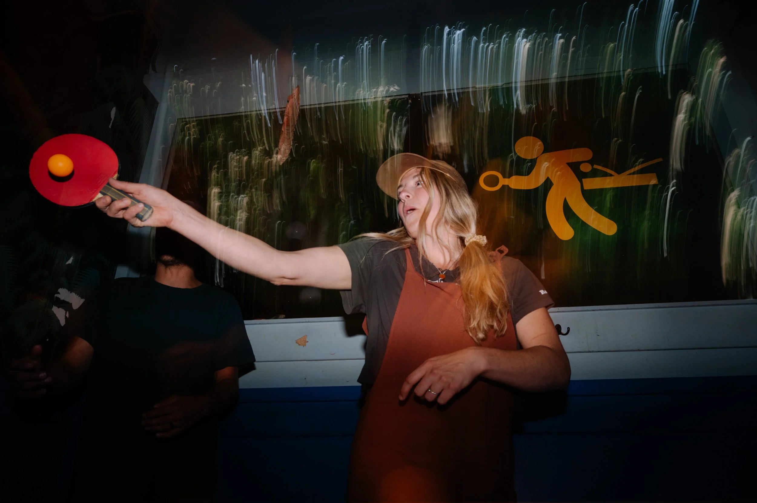 A woman with long red hair (possibly a waitress) wearing a brown apron and baseball cap, playing ping pong with a red paddle, in a dimly lit environment with blurred lights and a yellow sign of a person diving or performing a stunt in the background.