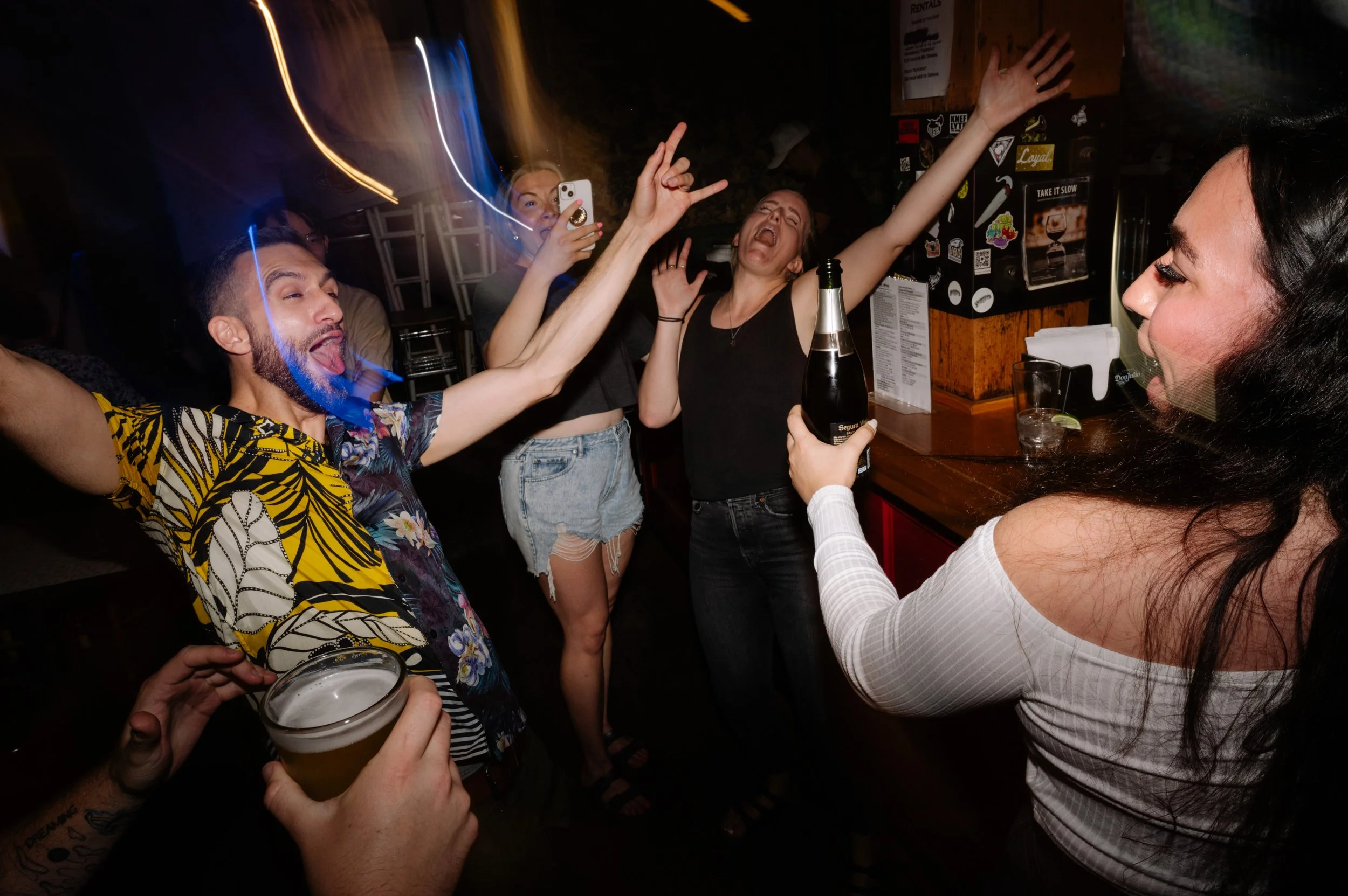 Group of friends dancing and celebrating in a bar, holding drinks, with colorful lights and a wooden bar counter in the background.