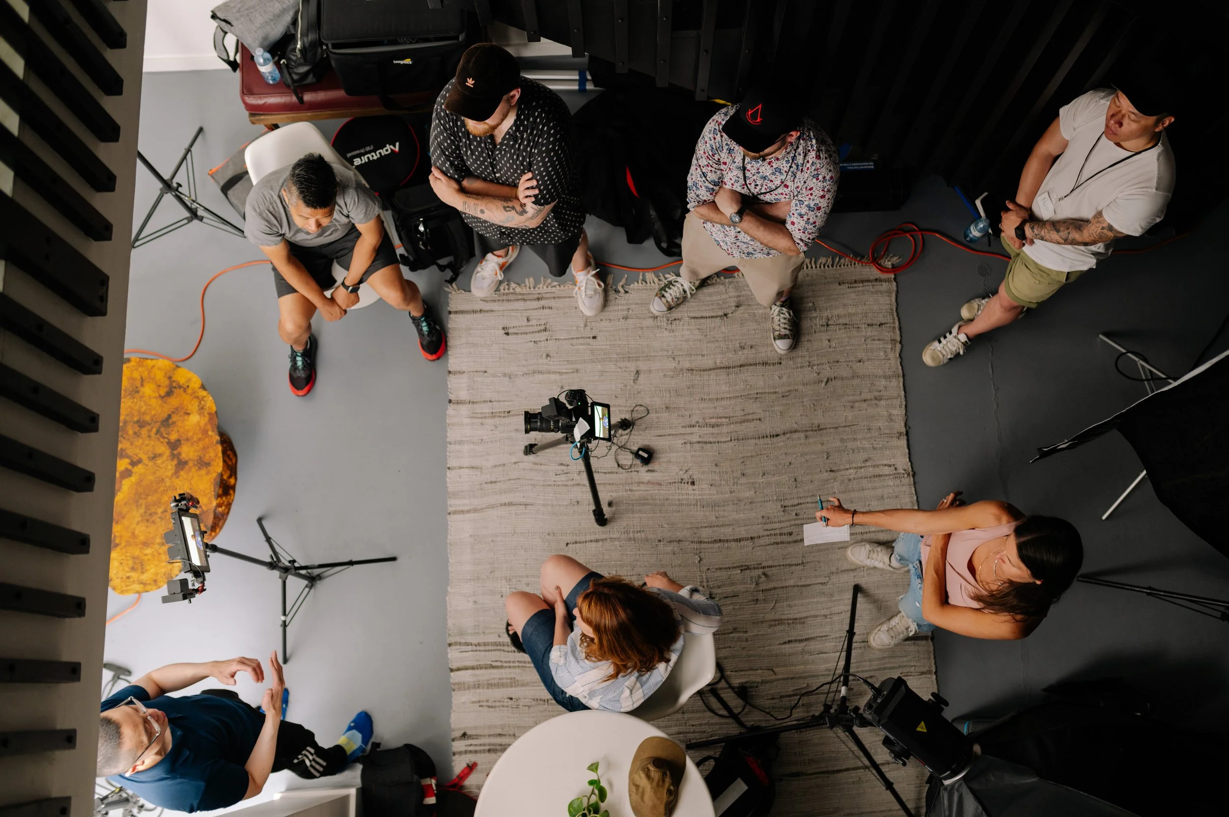 Overhead view of a group of people in a recording studio or podcast room. Five individuals are present, with one woman seated and the others standing or sitting around her. Cameras, tripods, and lighting equipment are visible, and the space includes 