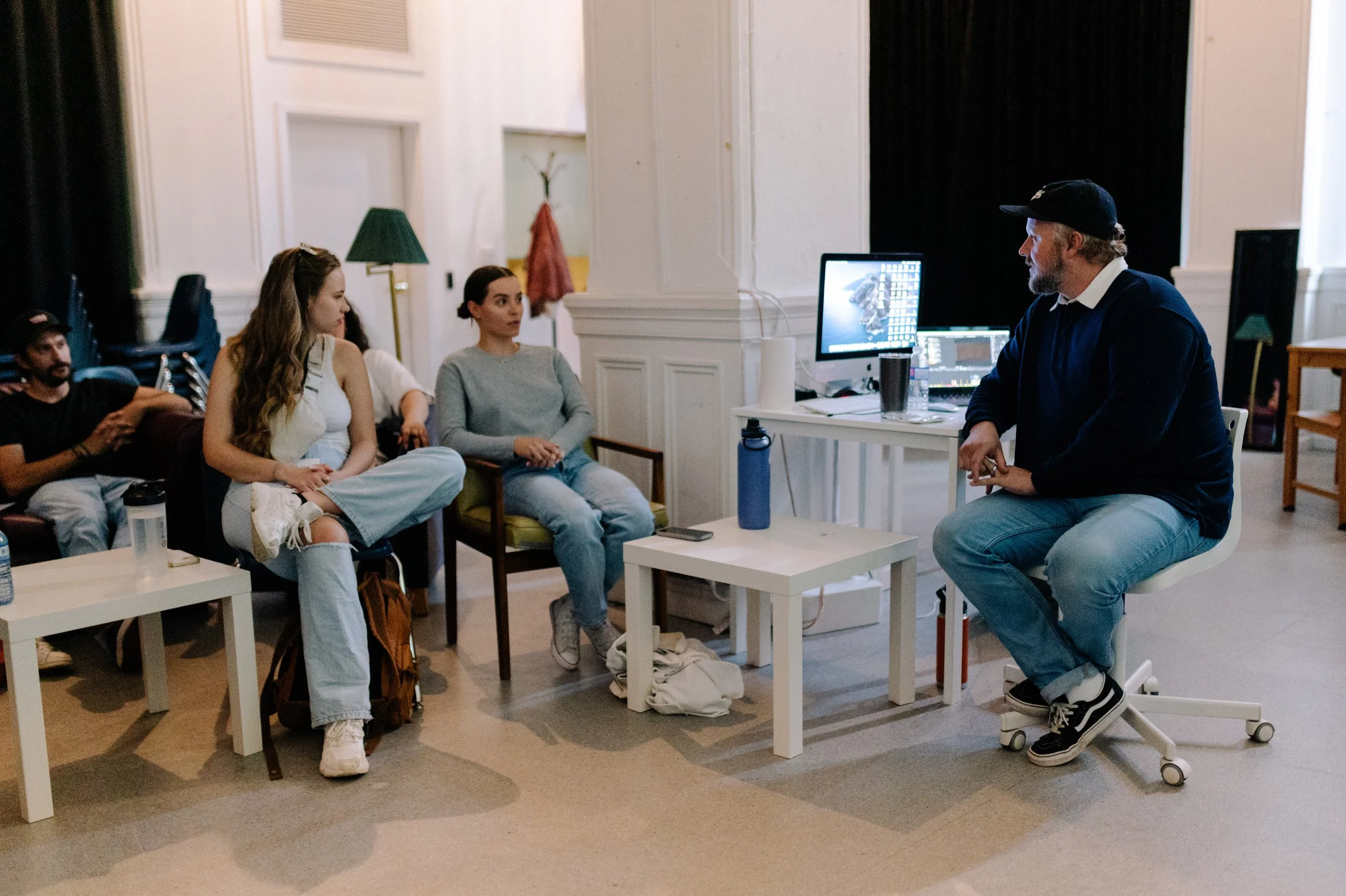A group of people sitting in a room with a man sitting on a rolling chair. The man has a beard, is wearing a cap, navy sweater, and jeans, and is facing the group. There are two women in front of him, one wearing a white dress and the other in a gray