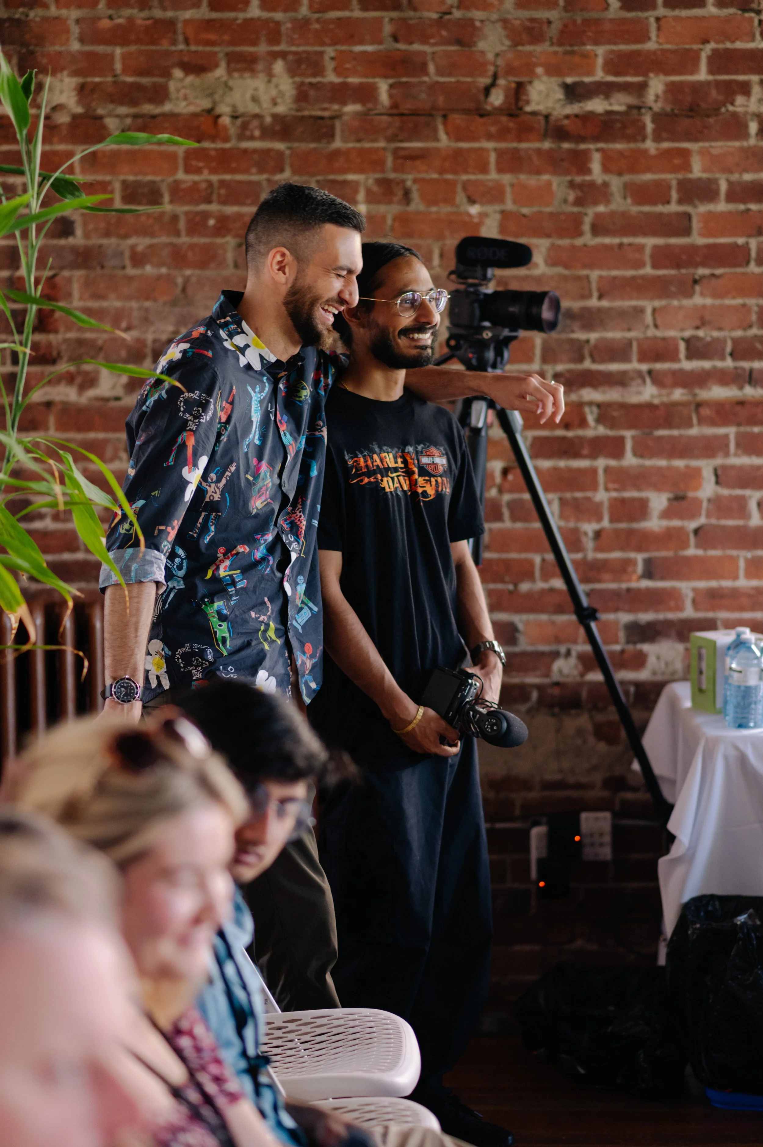Two men stand smiling near a brick wall, with one holding a camera and a third person filming with a camera mounted on a tripod behind them. In the foreground, blurred people are seated, with a table on the right holding water bottles and boxes.