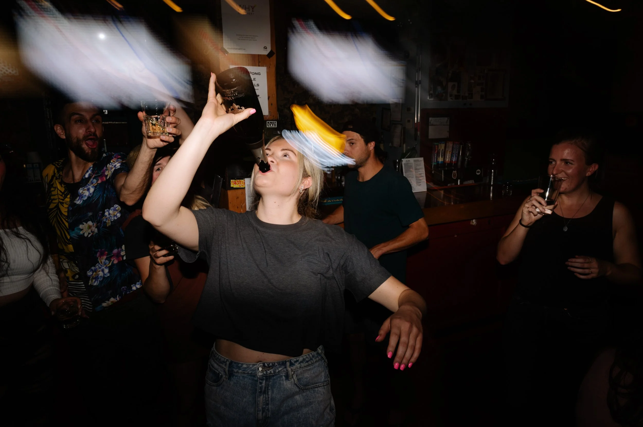 Group of people at a bar celebrating, with a woman in the center pouring a drink from a bottle into her mouth, surrounded by friends holding drinks and smiling.
