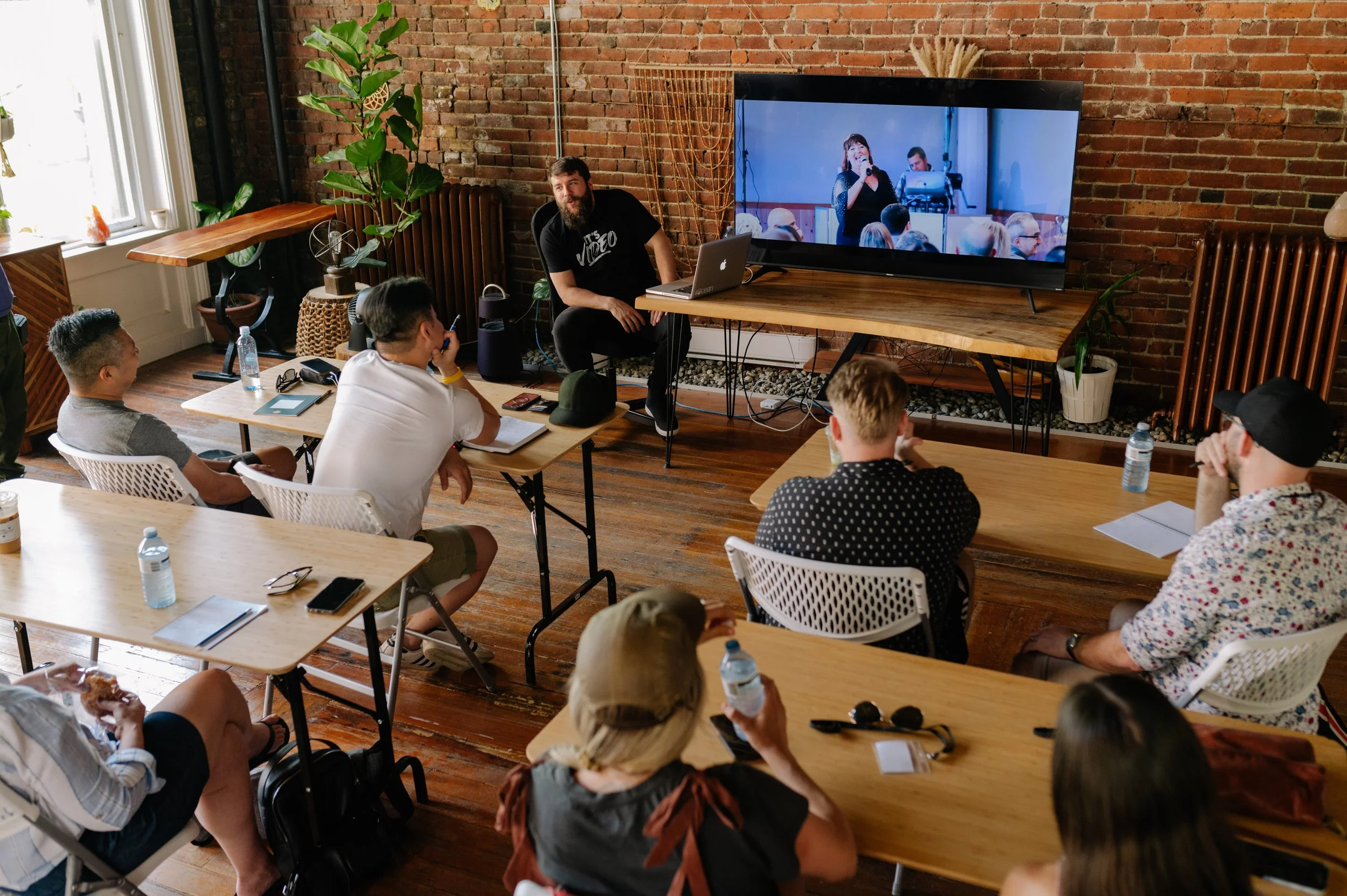 A group of people attending a presentation or workshop in a room with brick walls, seated at wooden tables with water bottles, notebooks, and electronic devices. The presenter, a bearded man wearing a black T-shirt, is sitting near a large screen dis
