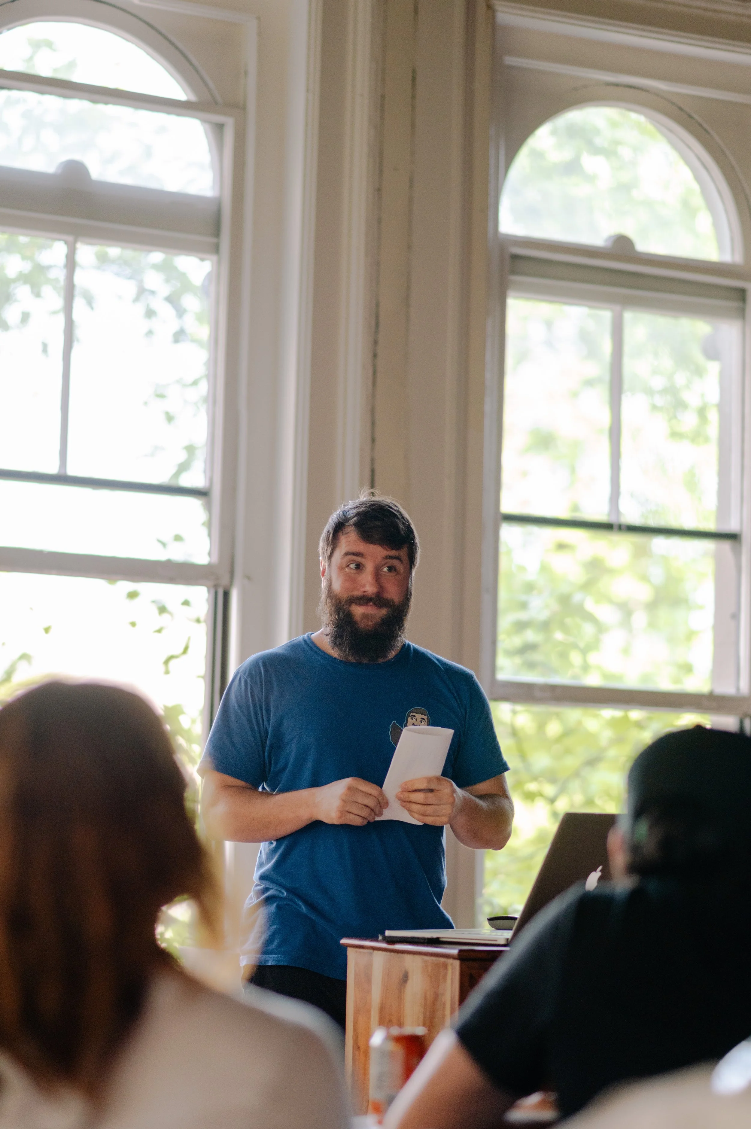Man with a beard holding a paper standing in front of a group, giving a presentation in a bright room with large windows.
