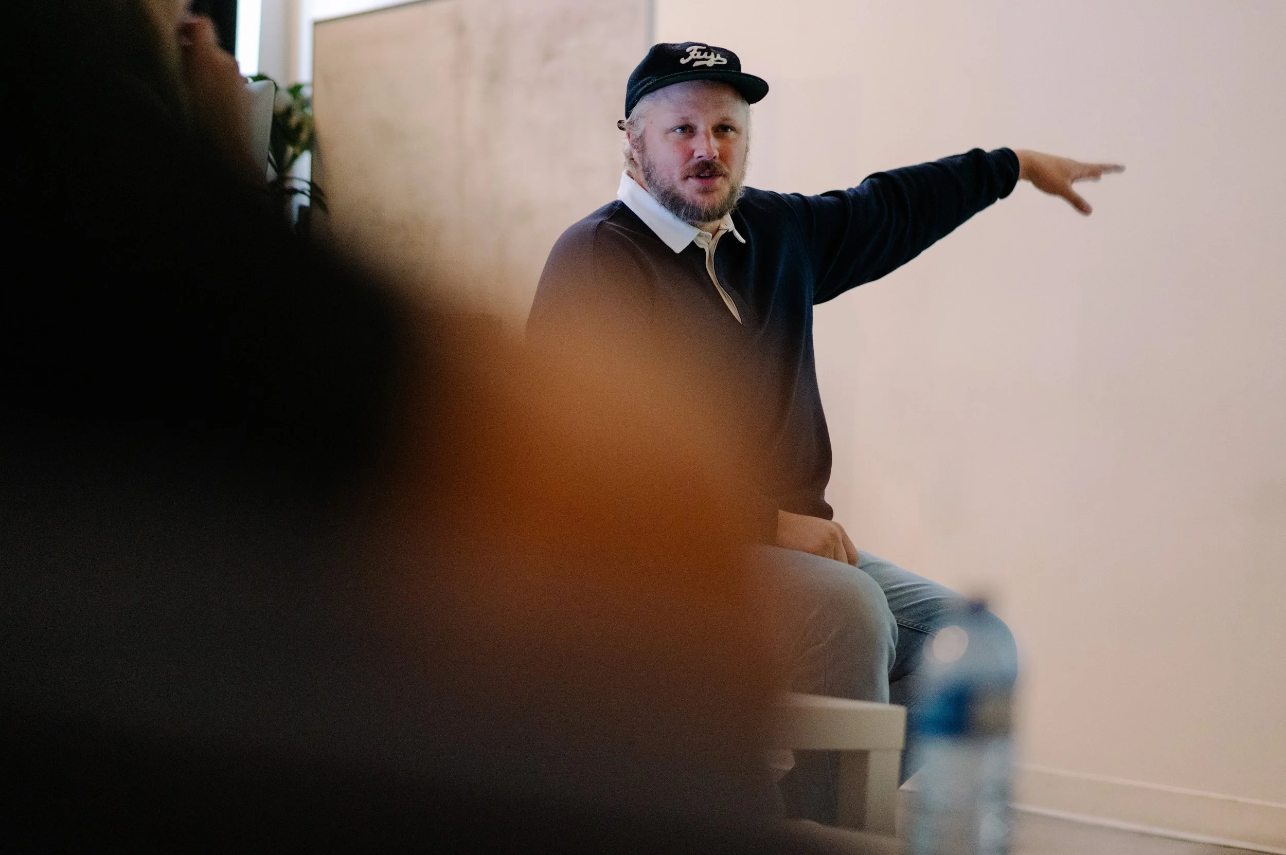 A man wearing a black baseball cap, black sweater, and light-colored pants is sitting on a bench, gesturing to the right with his arm extended, in a room with plain light-colored walls.