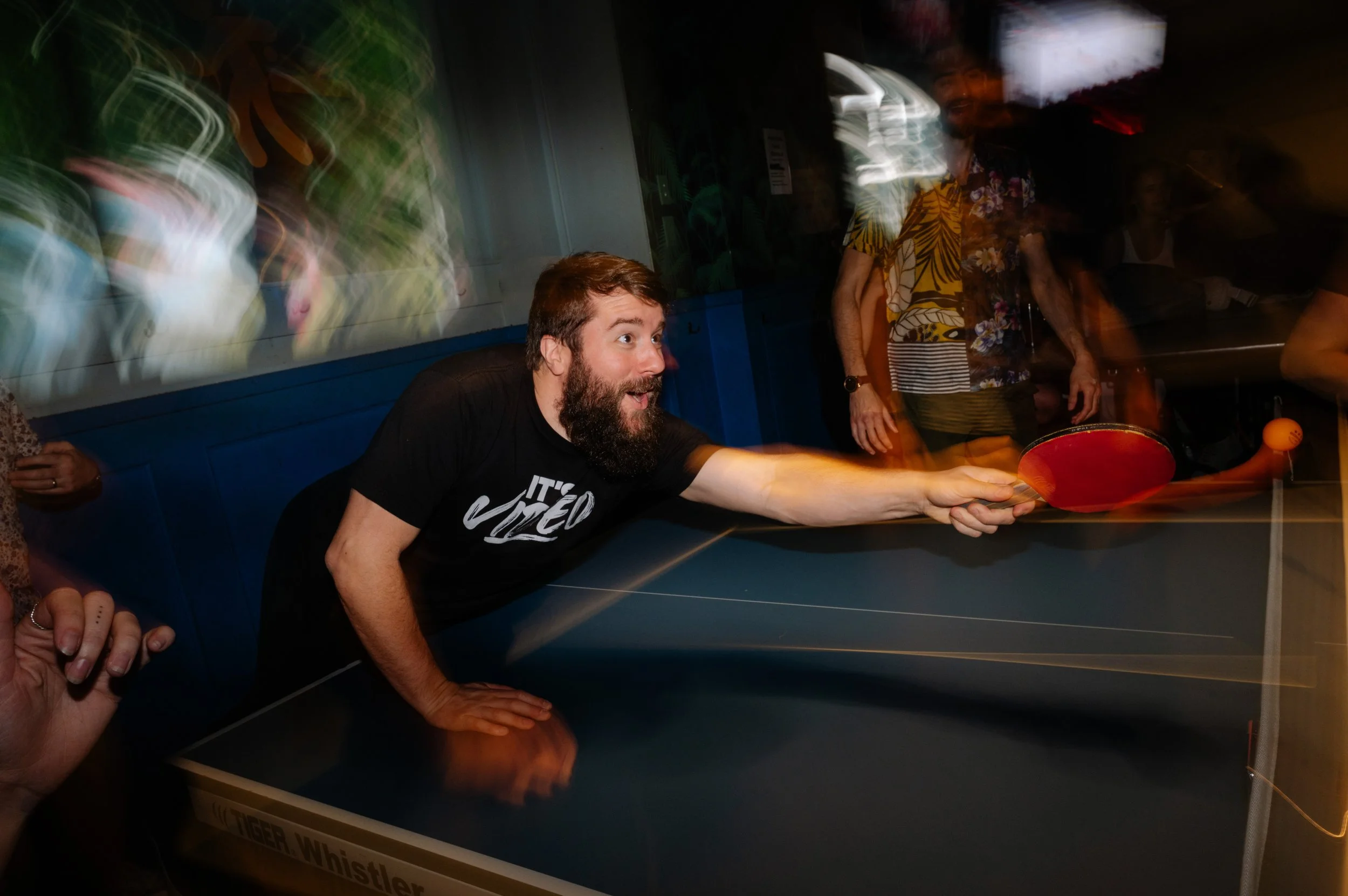 Man with a beard playing ping pong at a social gathering in a dimly lit room with colorful lights and people in Hawaiian shirts in the background.