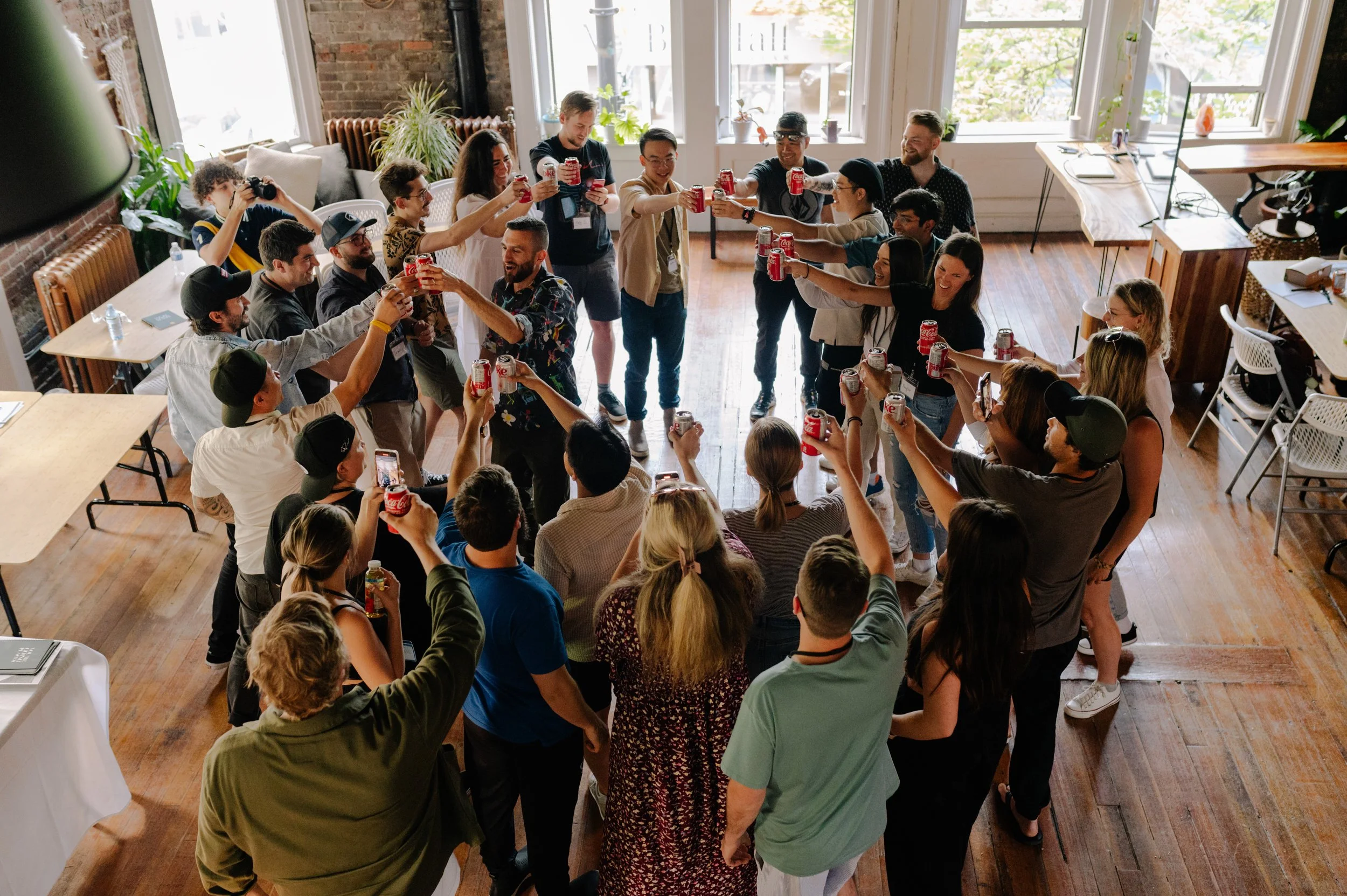 Group of people celebrating indoors, holding cans of soda and clinking them together in a circle.