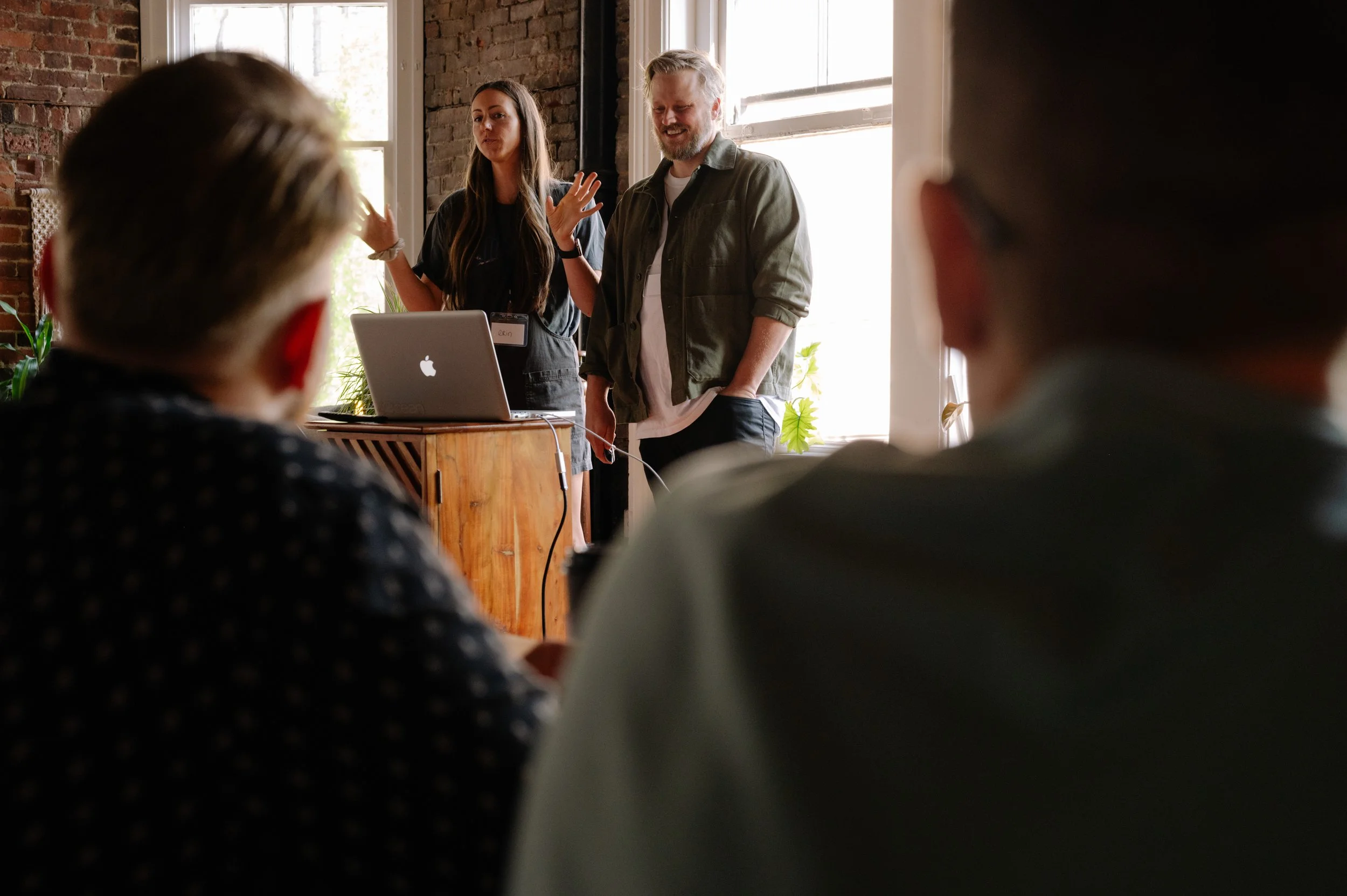 A woman is giving a presentation in a room with exposed brick walls, standing behind a wooden table with a laptop. She is gesturing with her hands, while a man stands beside her, smiling. Several people are seated, facing the presentation, with their