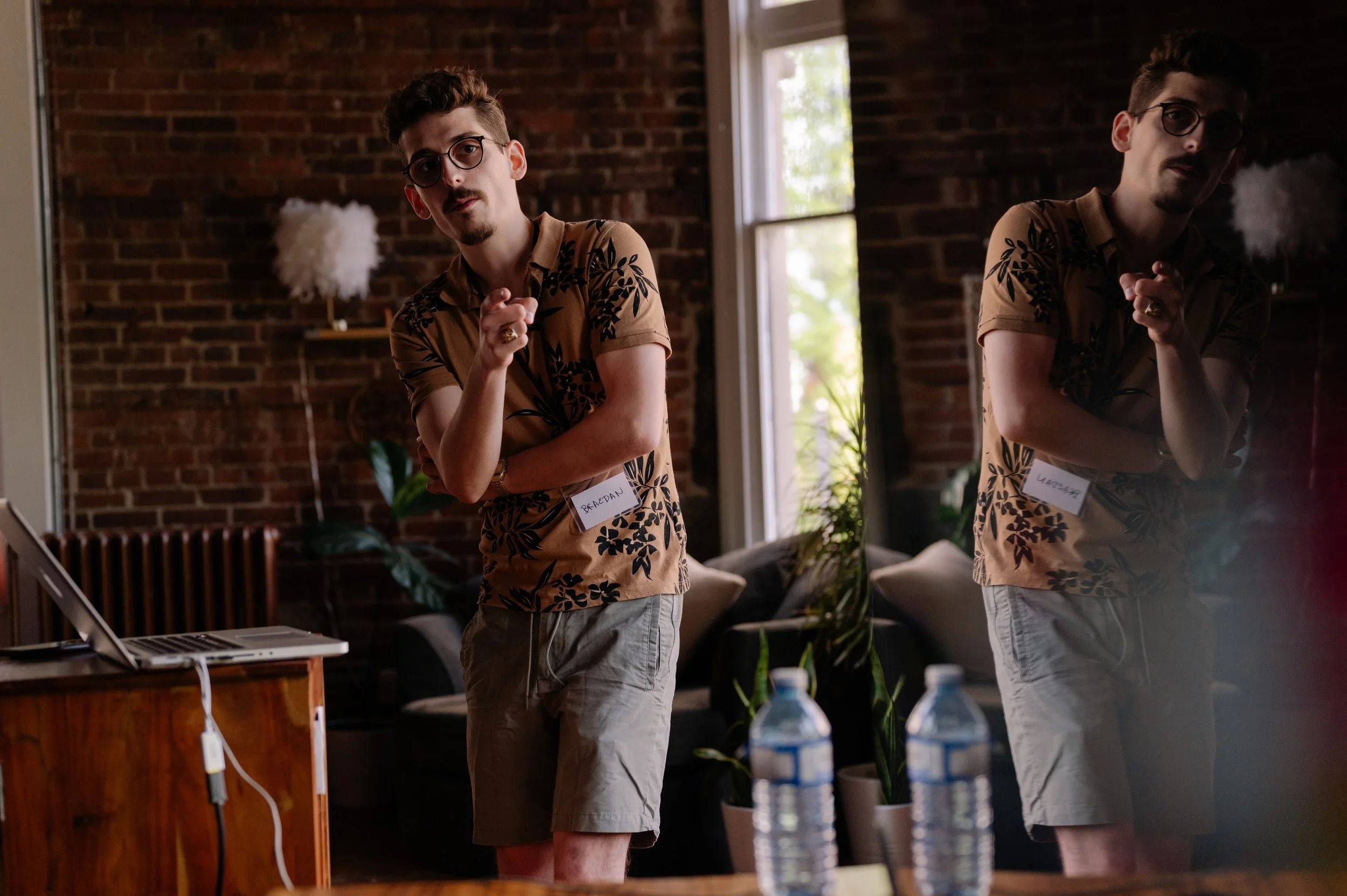 Young man with glasses and a mustache, wearing a tan shirt with black floral pattern and gray shorts, pointing towards the camera in a cozy room with brick wall, plants, and natural light.