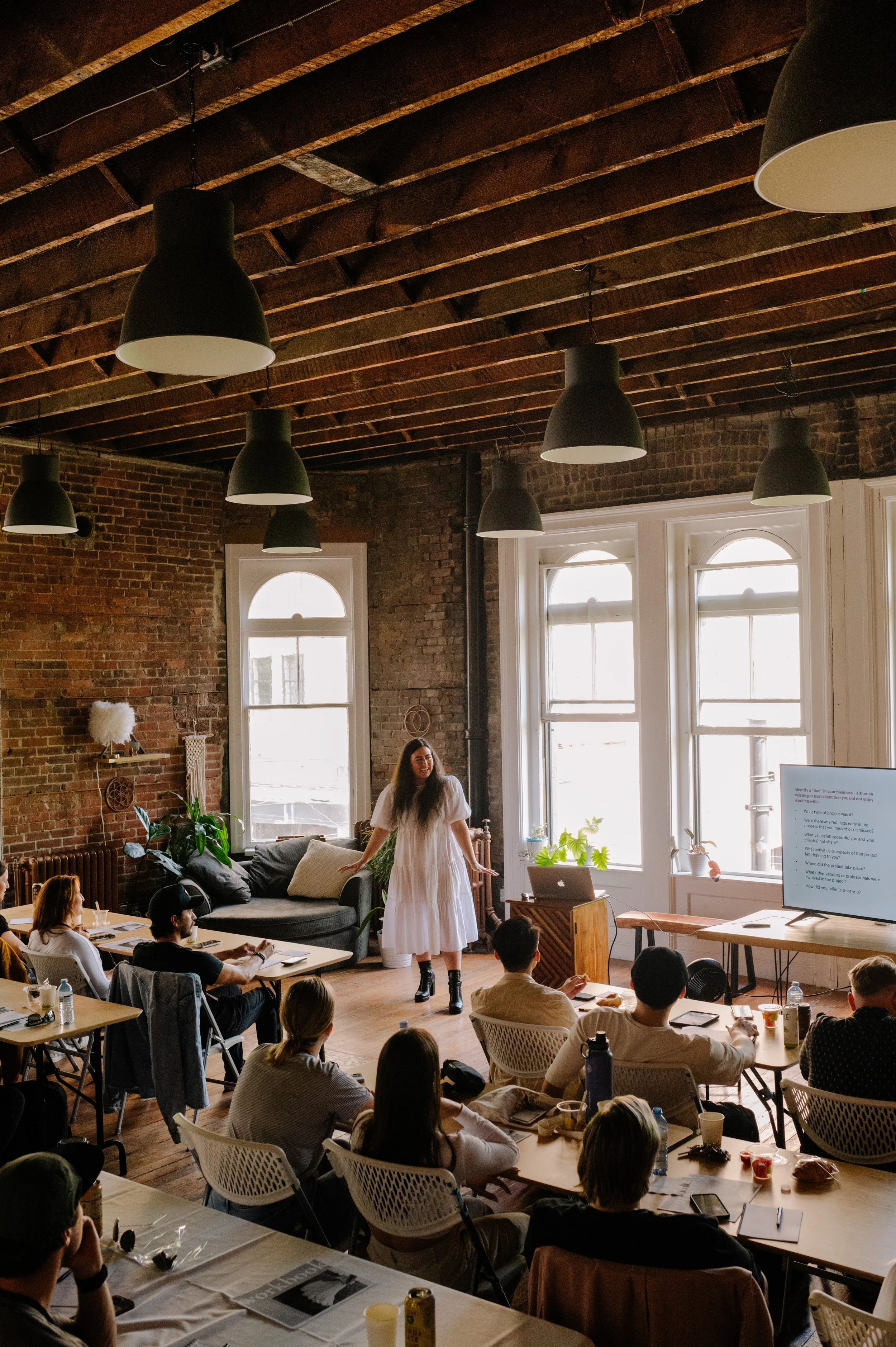 A woman gives a presentation to a group in a rustic, brick-walled classroom with large windows, black pendant lights, and a television screen.