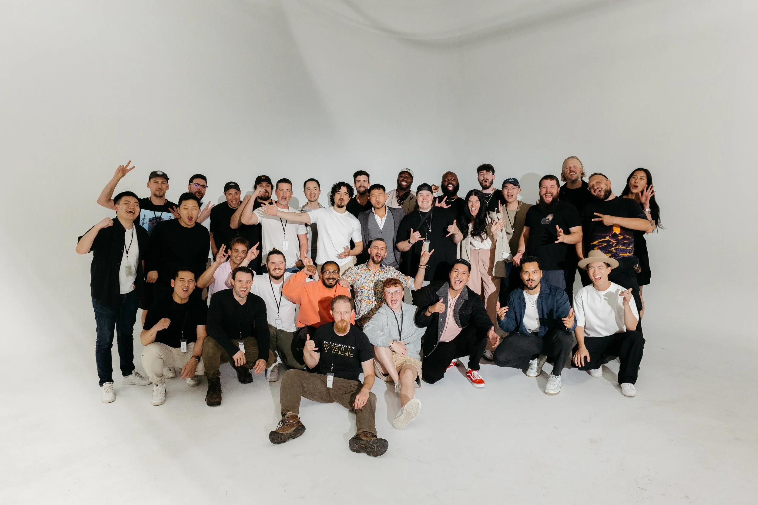 A large group of diverse people posing together for a photo in a studio with a white background, smiling and making various hand gestures.