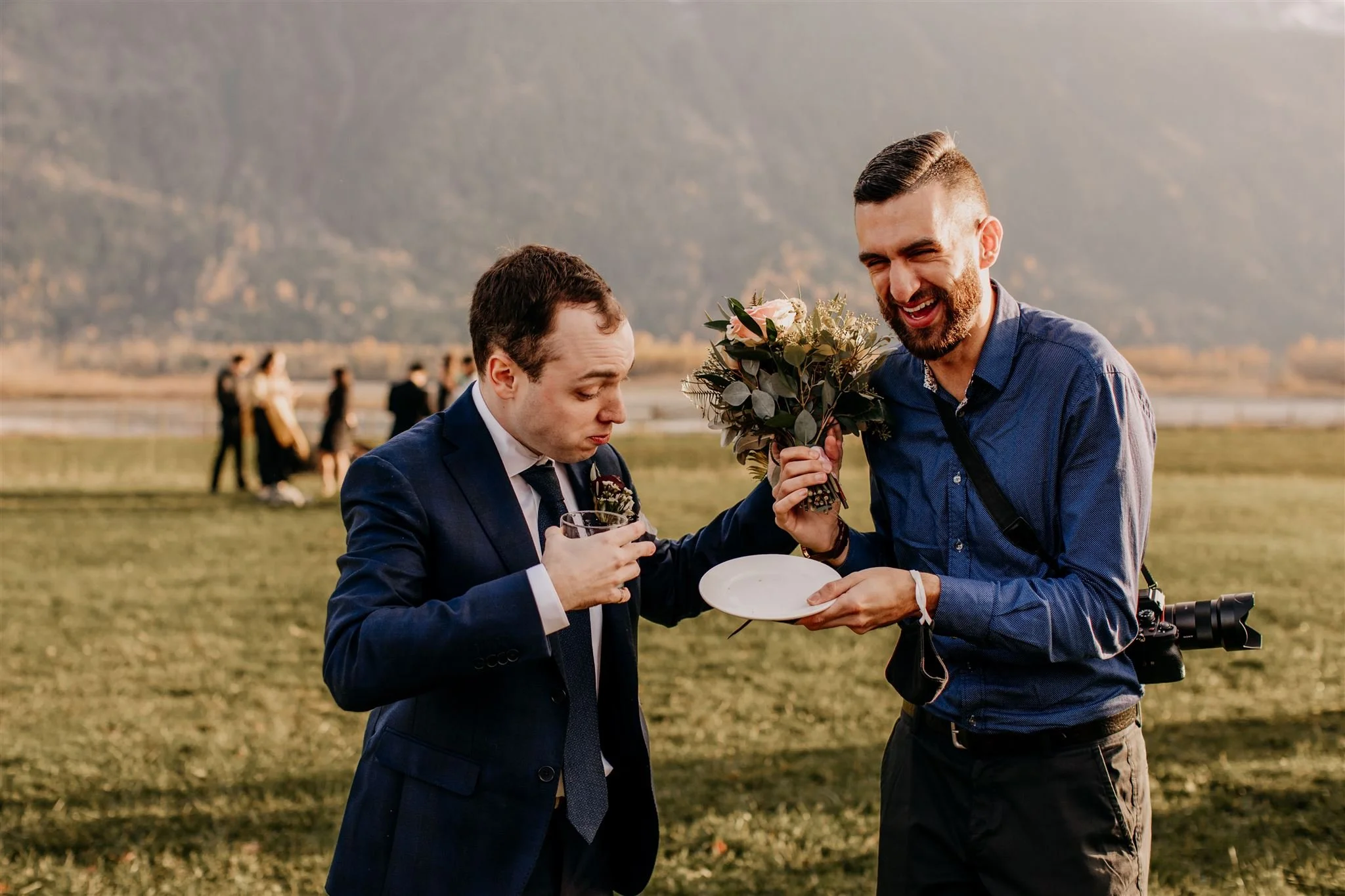 Two men at an outdoor event, one in a suit holding a drink and the other in casual clothing with a camera, sharing a bouquet and a plate, smiling with mountains in the background.