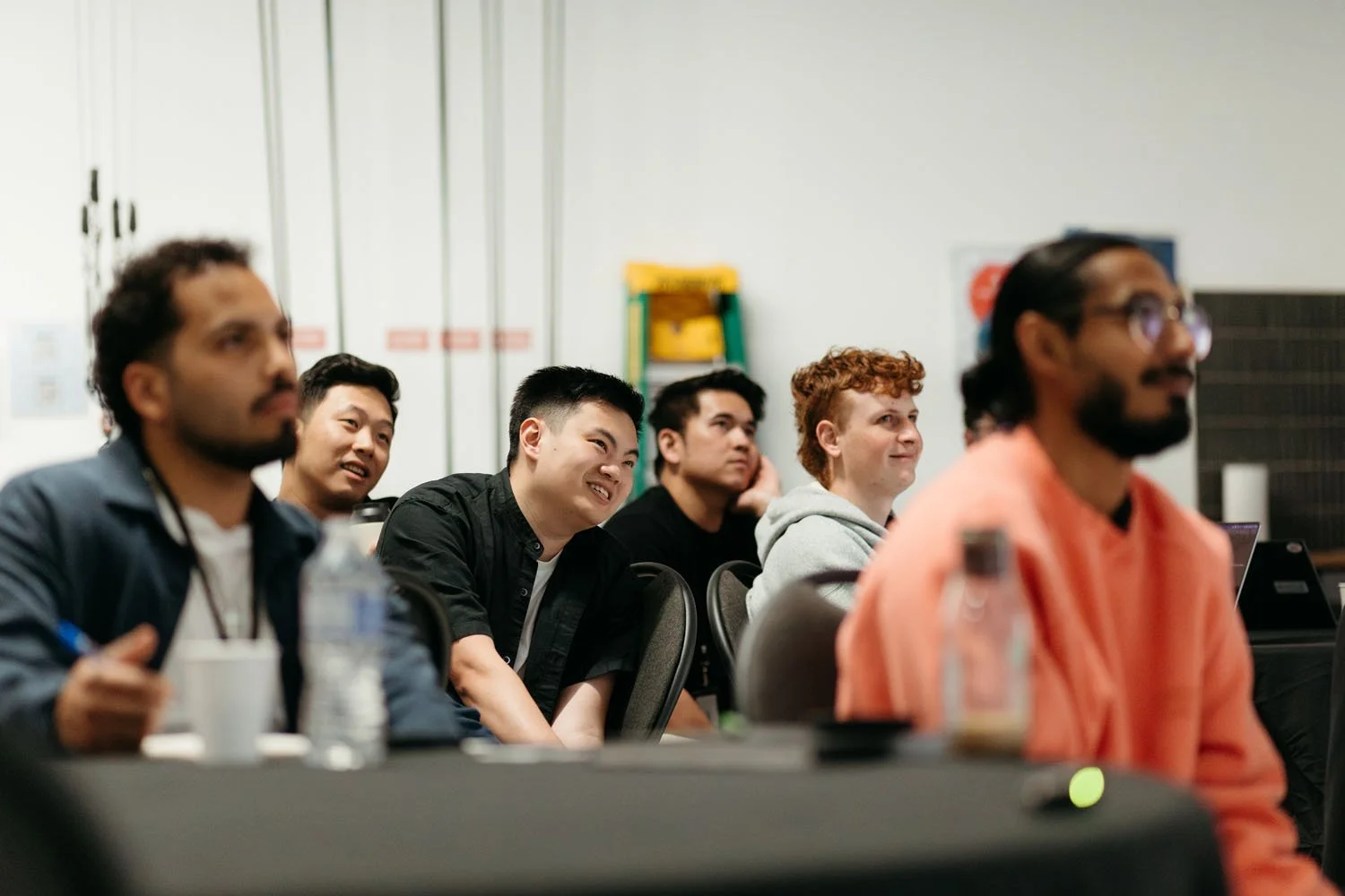 A diverse group of six young adults sitting in a classroom or conference setting, attentively listening and smiling, with equipment and bottles on the table in front of them.