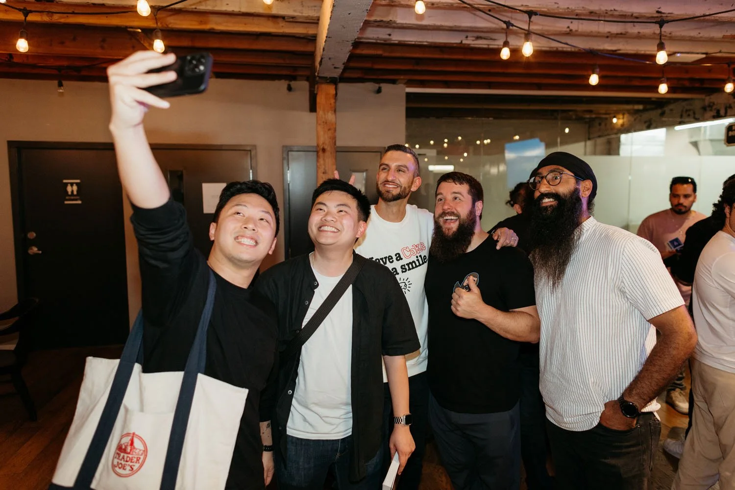 Group of five men taking a selfie together inside a restaurant or cafe. The men are smiling and appear to be enjoying themselves.