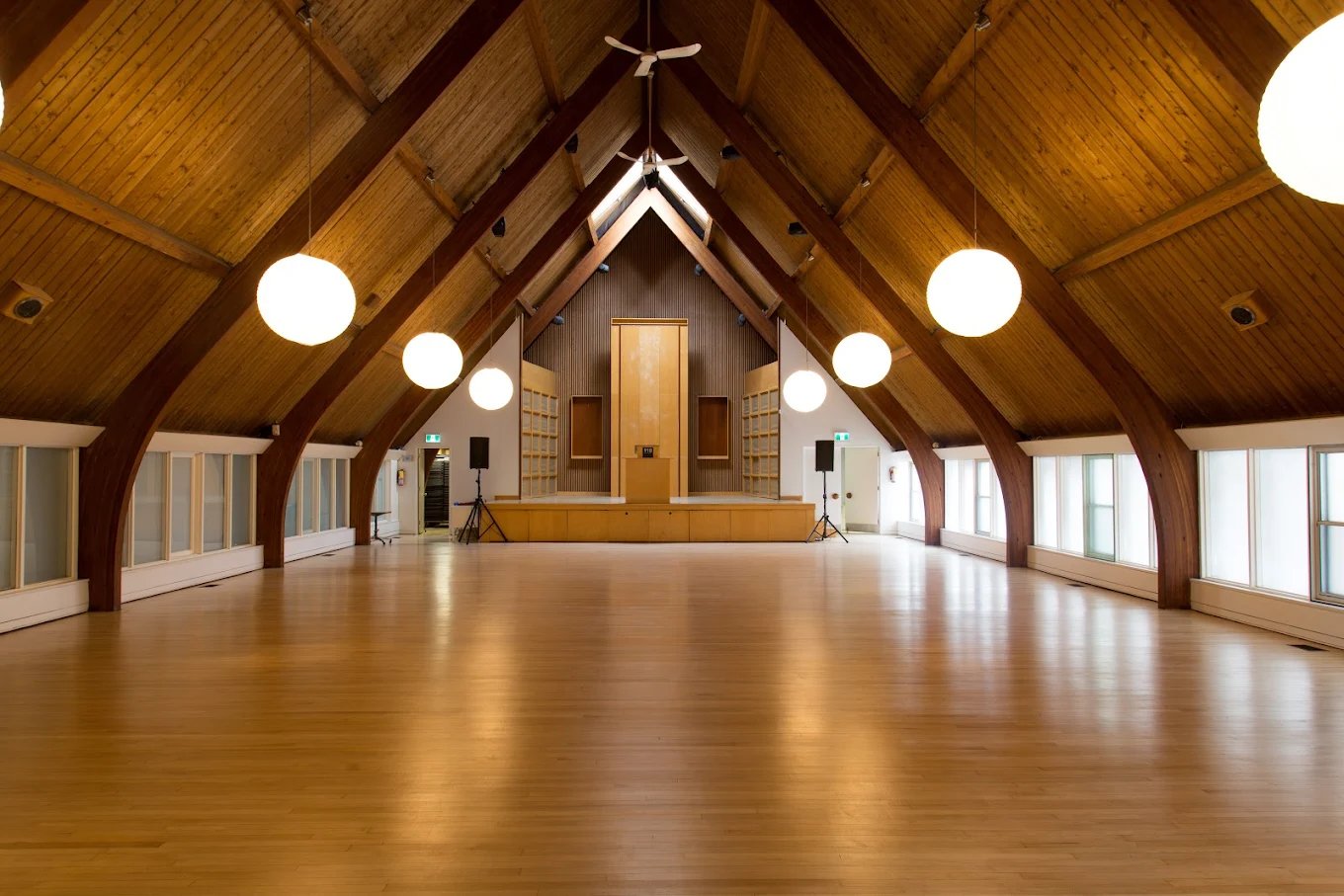 Empty wooden hall or auditorium with a stage at the front and hanging round lights from the arched wooden ceiling.