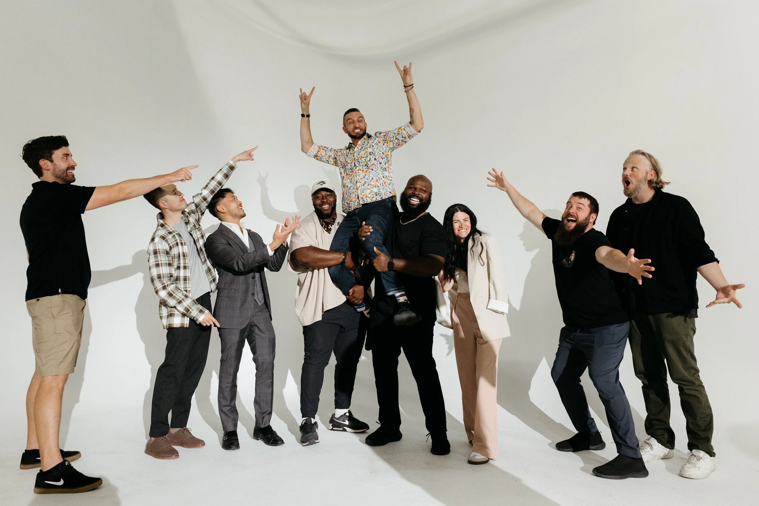 A diverse group of nine happy people celebrating, with one man being lifted in the air and others cheering, in a plain white studio