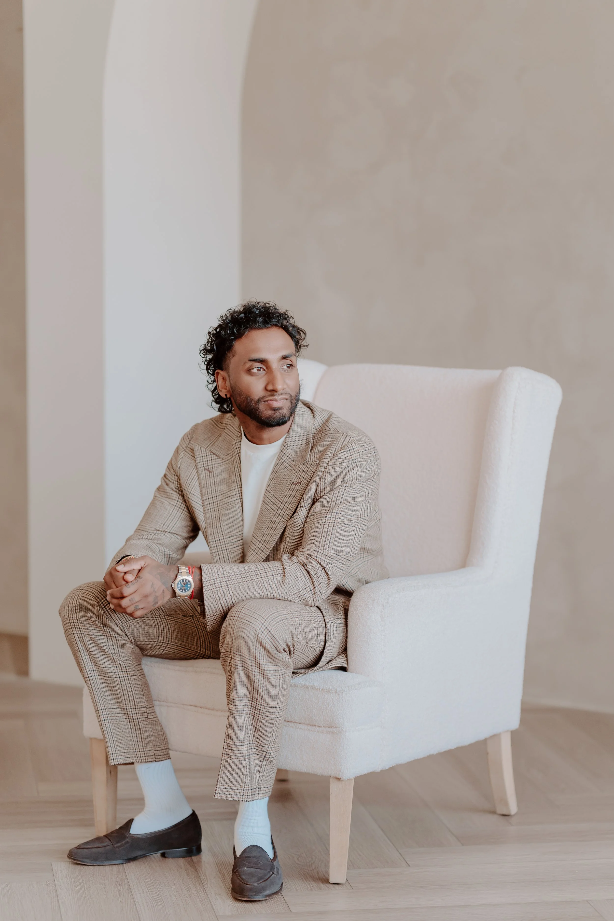 A man with medium black hair in a beige pin stripe suit sitting on a white chair against a cream backdrop.