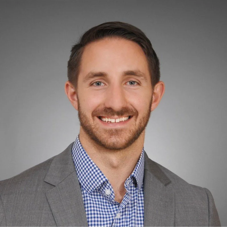 Headshot of a young man with short brown hair, light beard, wearing a gray suit jacket and blue checkered shirt, smiling against a gray background.