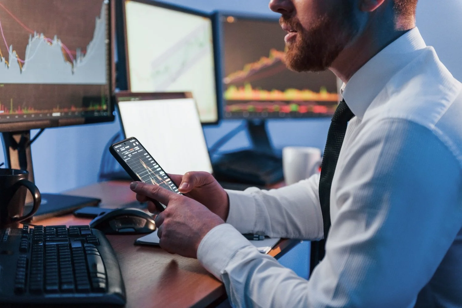 Man in white shirt and tie analyzing stock trading charts on smartphone with multiple monitors displaying financial data and graphs in background.