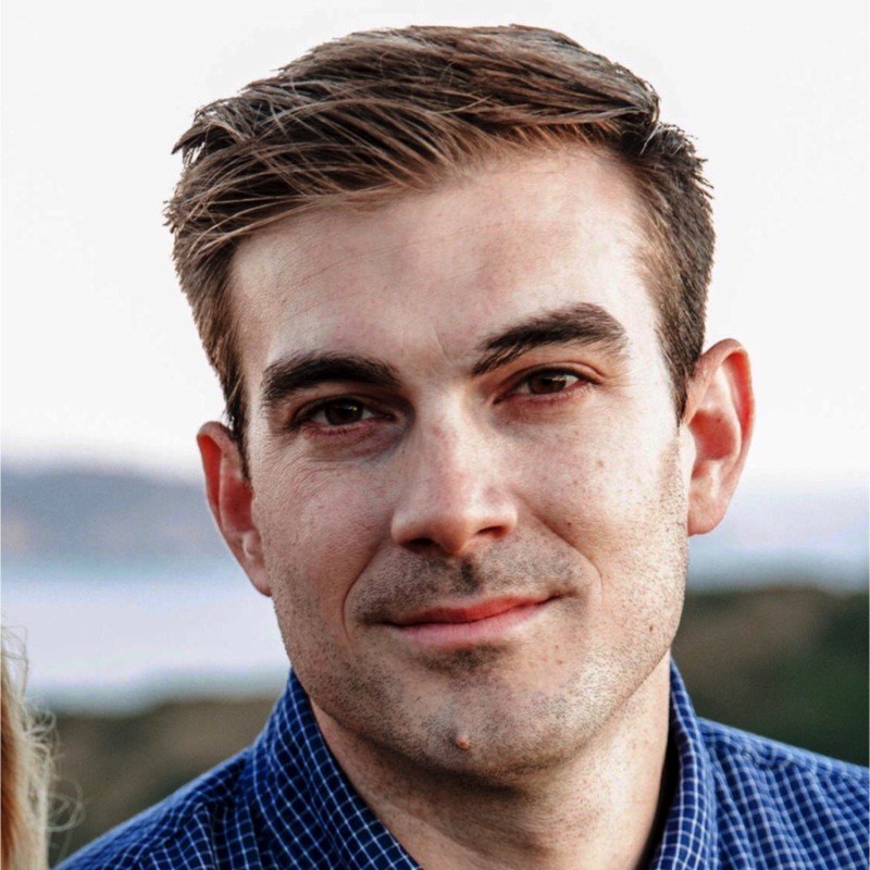 Close-up photo of a young man with light brown hair, wearing a blue checkered shirt, outdoors with a blurred background.