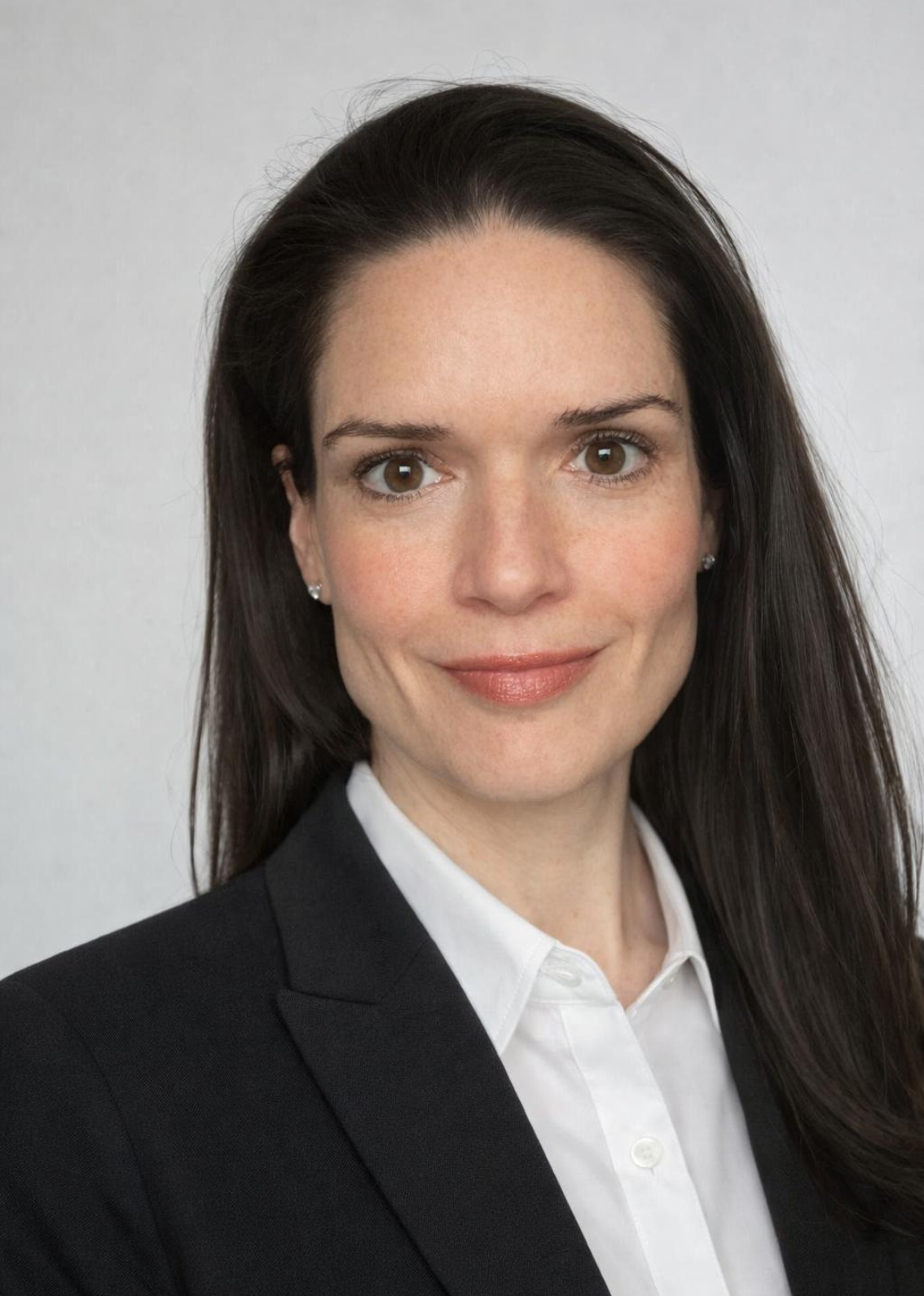 A professional woman with long dark hair, wearing a black blazer over a white shirt, is smiling and looking directly at the camera against a plain neutral background.