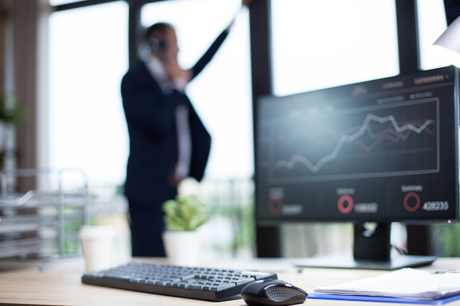 Office desk with computer monitor displaying a line graph, keyboard, mouse, and papers. Blurred woman in business attire talking on phone in background.