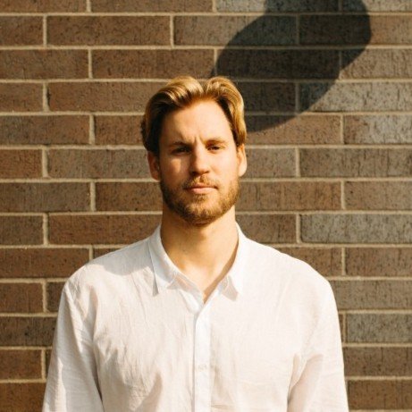 A man with light brown hair and a beard standing against a brick wall, wearing a white shirt with sunlight casting a shadow in the background.