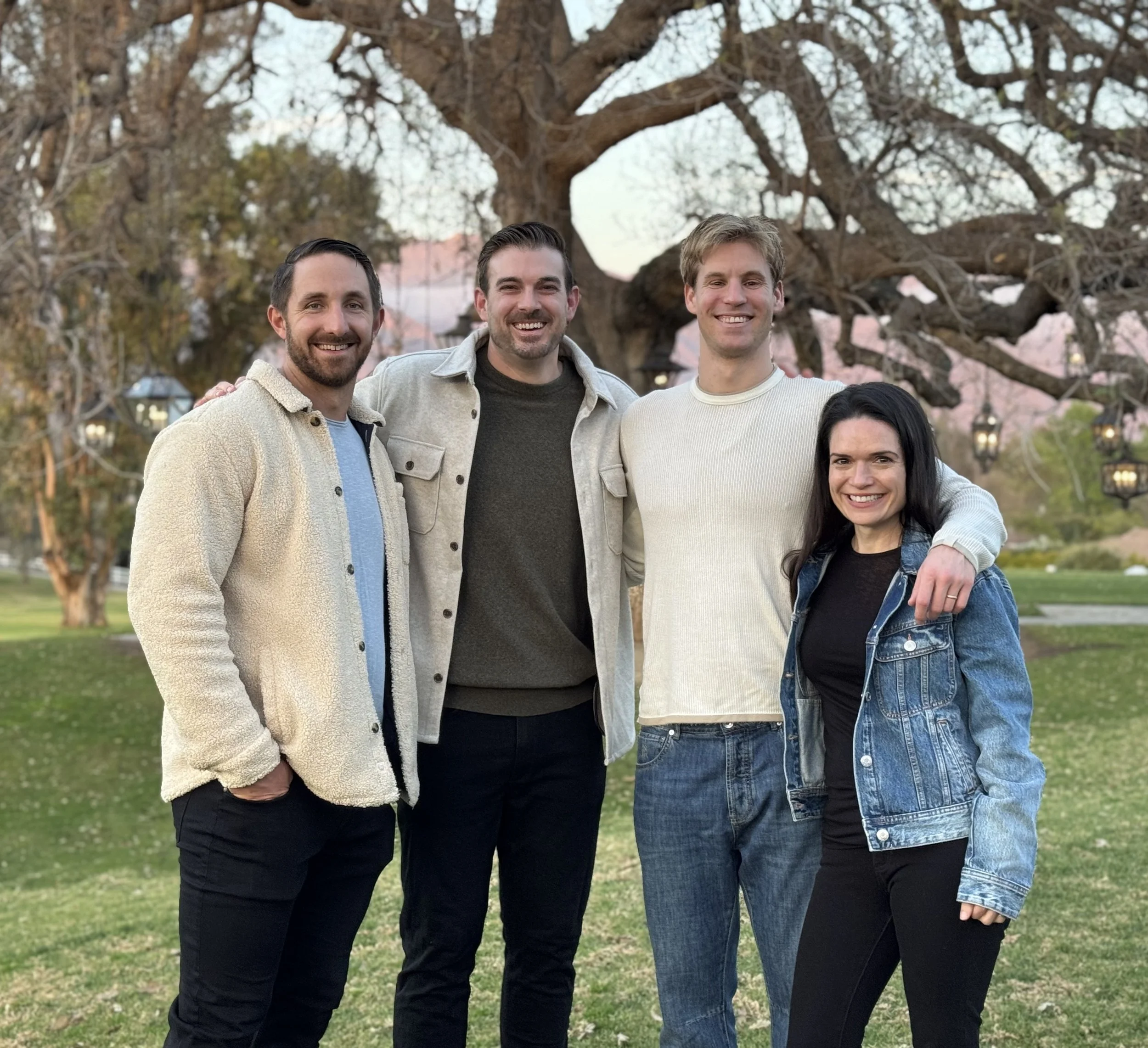 Group of four friends smiling and posing outdoors in front of a large tree in a park during spring.