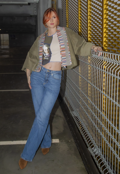 A woman with short red hair wearing a gray crop top, denim jeans, an oversized beige jacket, and a colorful striped scarf, standing in a parking garage next to a metal railing.
