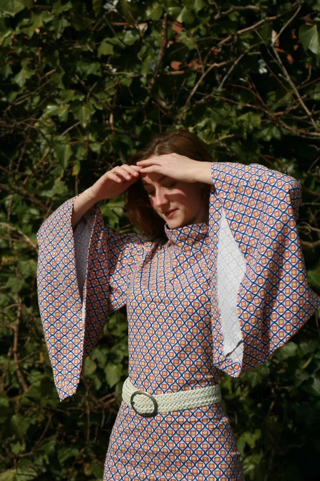 A woman with brown hair standing outdoors against a background of green leafy vines, wearing a colorful patterned dress with wide sleeves and a white woven belt, shielding her eyes from the sun with her hand.