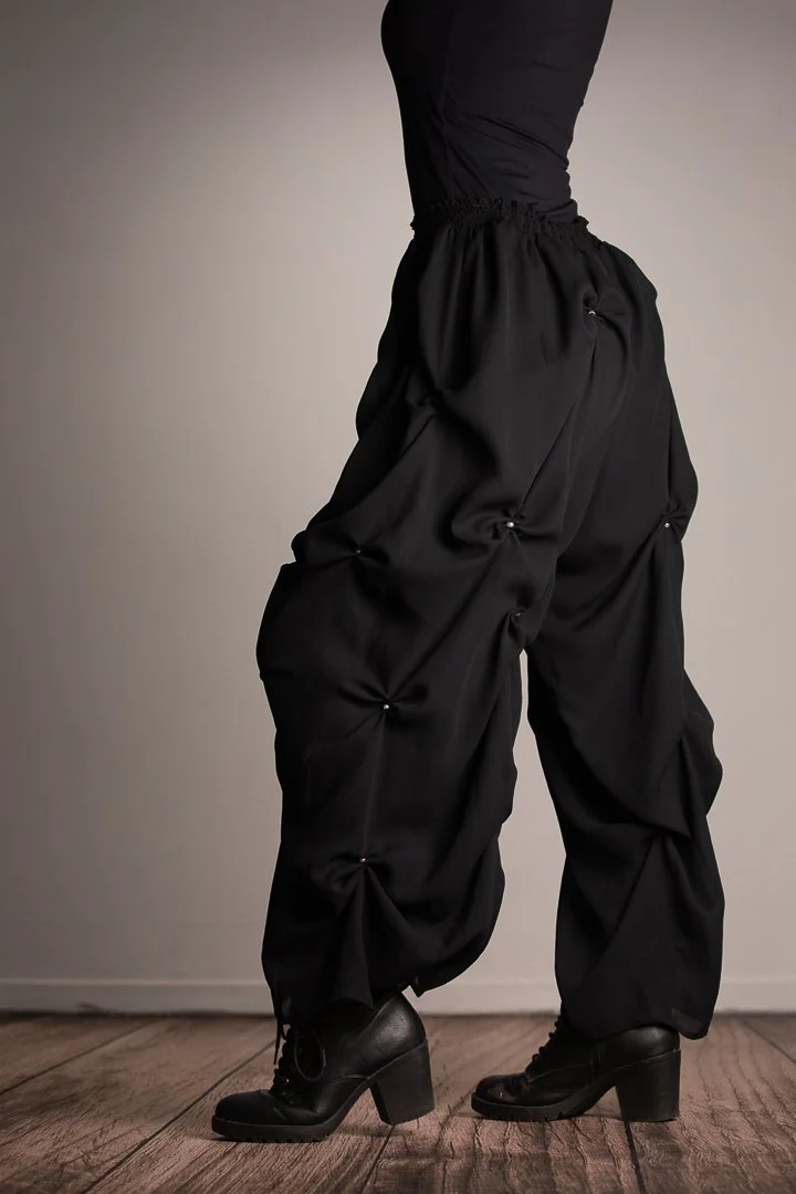 Close-up of a person wearing black cargo pants with ruched details and black heeled combat boots, standing on wooden floor against a plain wall.