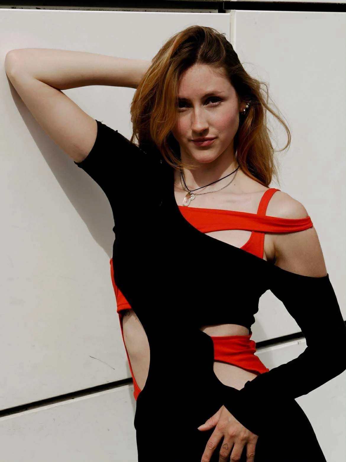 Young woman with red hair posing against a white wall, wearing a black and red dress with cutouts, and jewelry.
