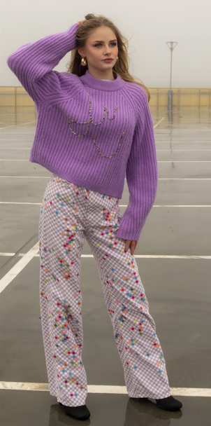 A young woman standing on a rooftop parking lot wearing a purple knitted sweater with the word 'love' written in gold, colorful patterned pants, and black shoes.
