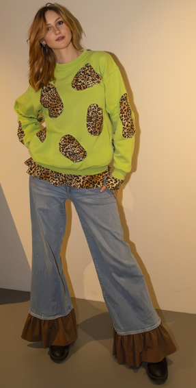 Woman wearing a lime green sweatshirt with leopard print patches, light blue wide-leg jeans, and brown ruffled shoes.