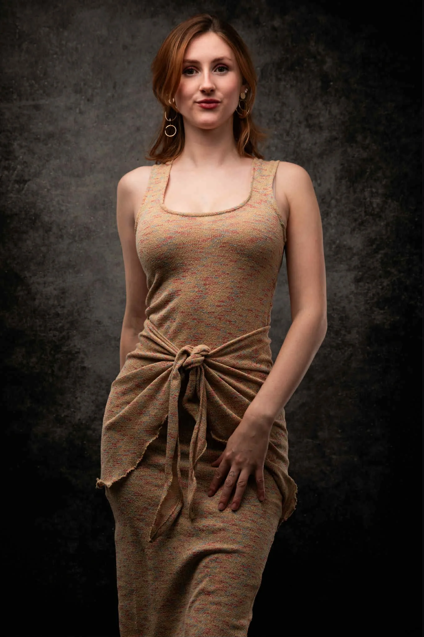 A woman with red hair wearing a sleeveless, multicolored, textured dress with a knot at the waist, standing against a dark textured background.