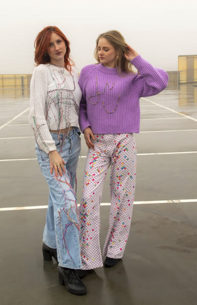 Two women standing on a rooftop parking garage, one with red hair wearing a white sweater and jeans, the other with blonde hair wearing a purple sweater and patterned pants.