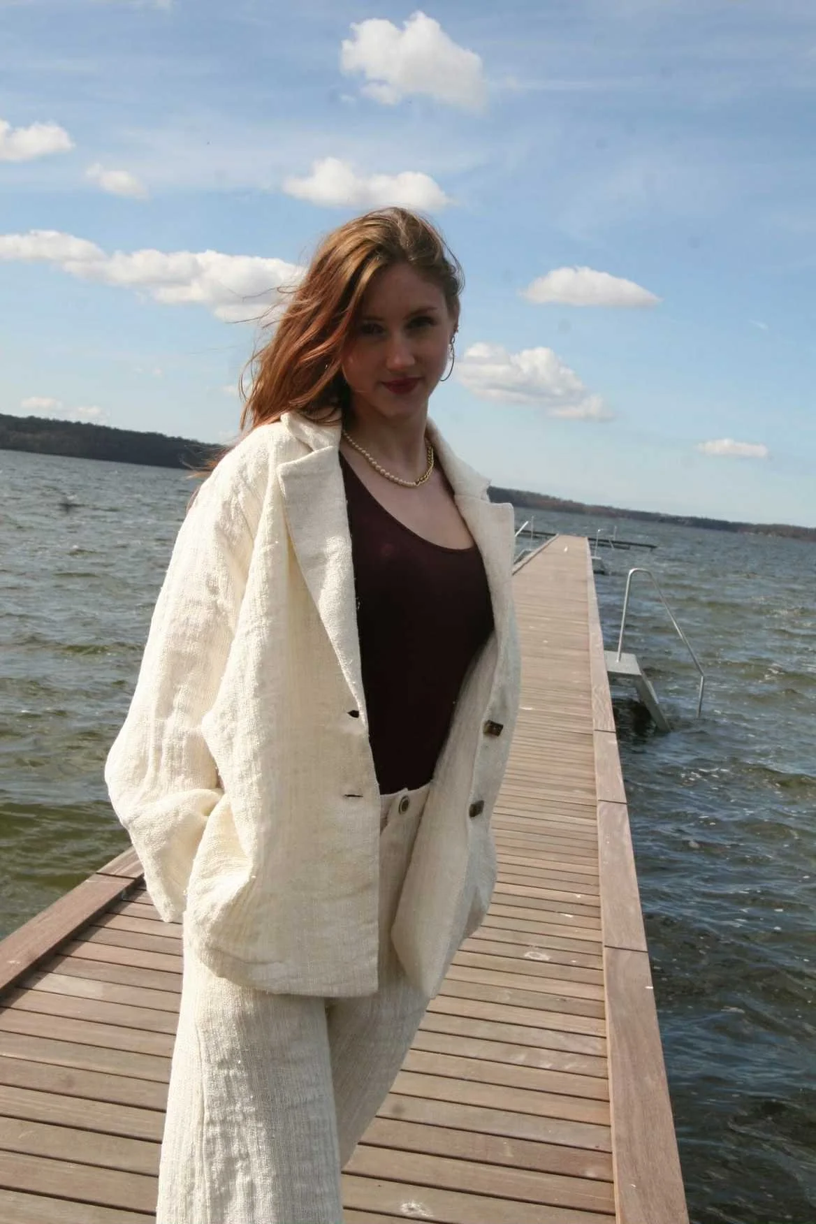 A woman standing on a wooden dock by the water under a partly cloudy sky, wearing a cream-colored suit with a black top and gold jewelry.