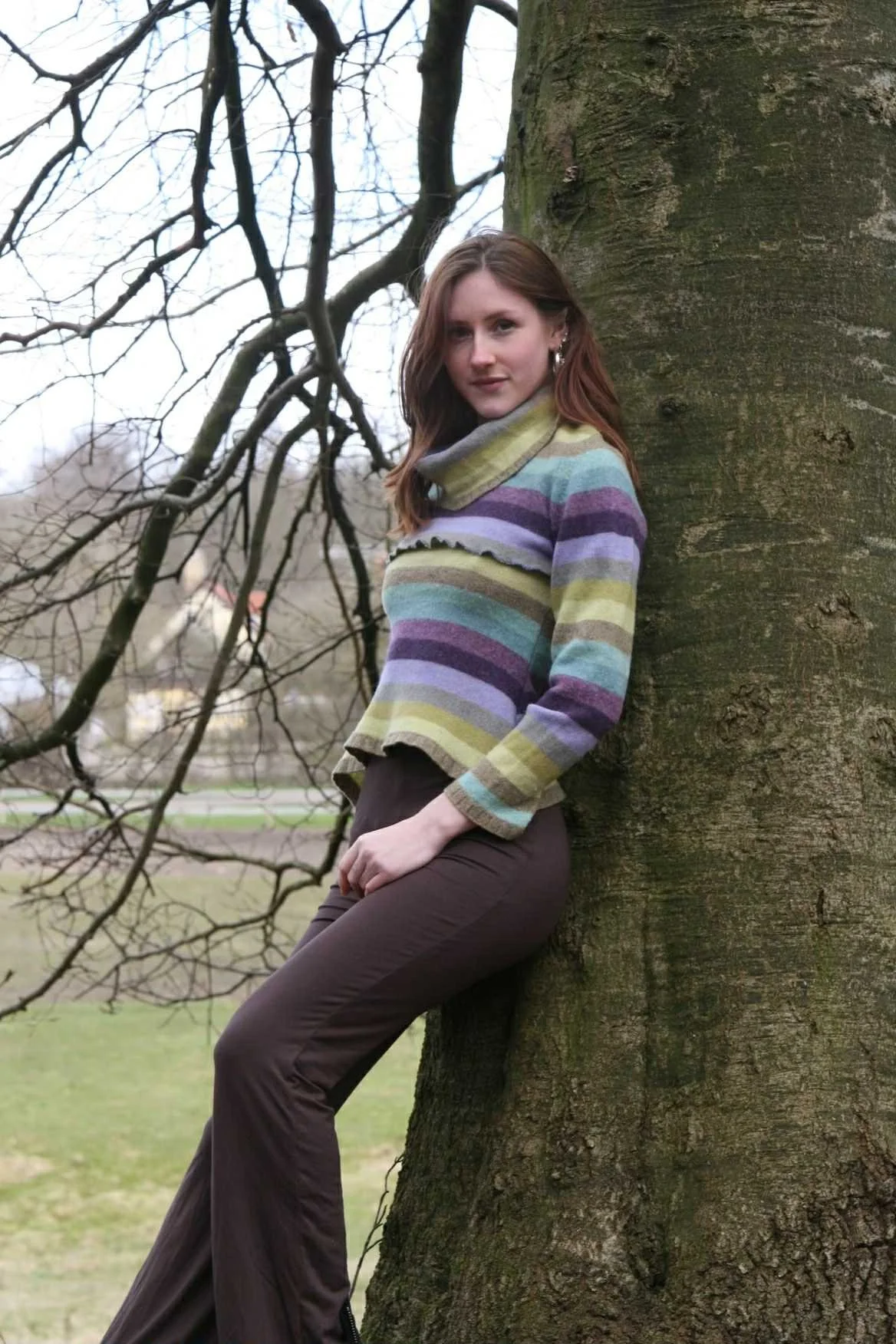 A young woman with brown hair leaning against a large tree trunk outdoors, wearing a colorful striped sweater and brown pants, with a background of leafless branches and a park.
