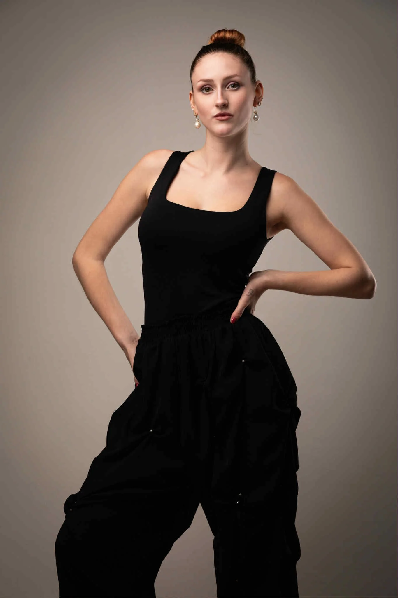 A young woman with red hair in a bun, wearing a black sleeveless top and black pants, standing against a plain gray background.