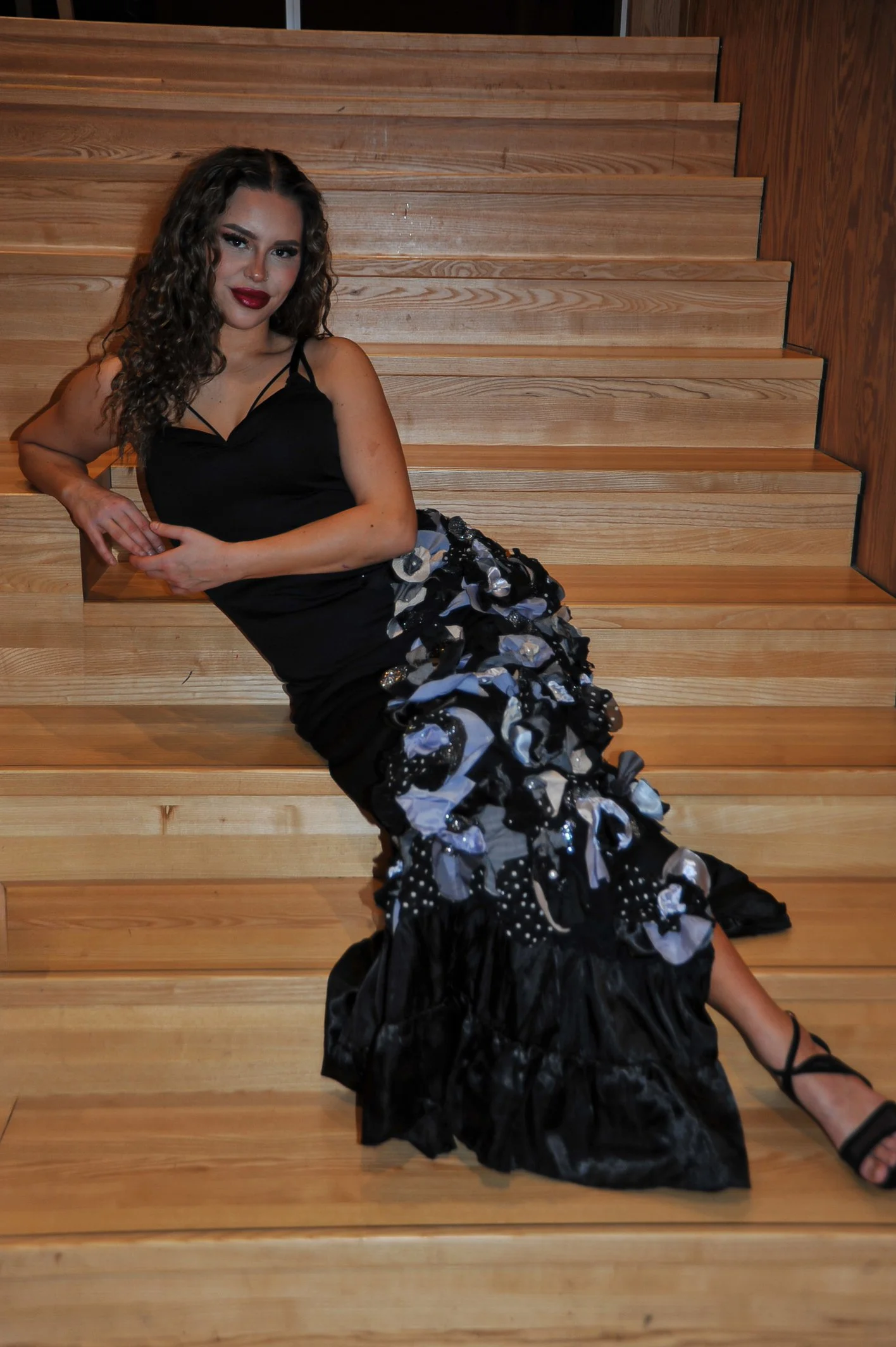 A woman with curly hair, wearing a black dress with spaghetti straps and a ruffled black and white skirt, is sitting on wooden stairs.