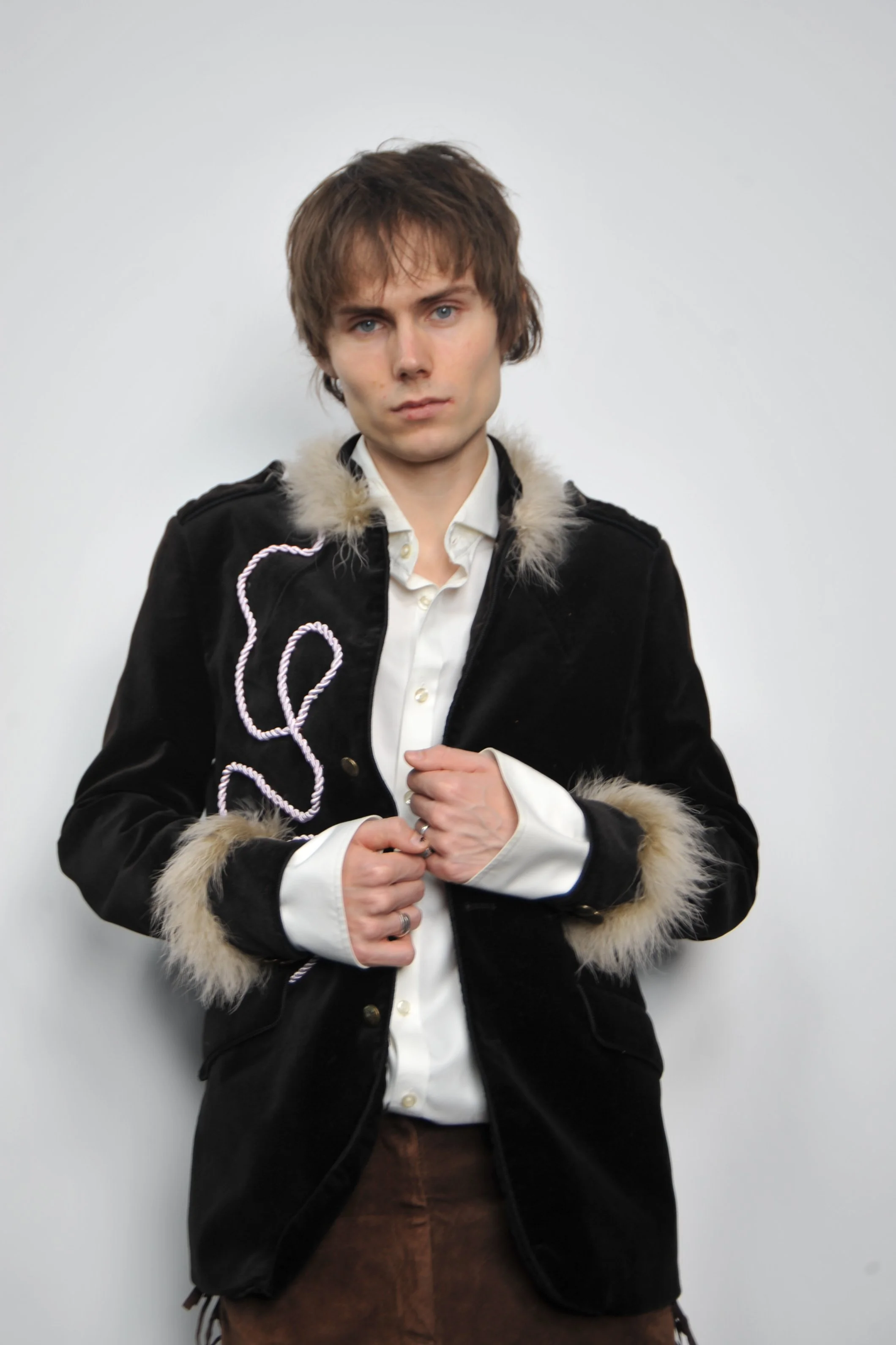 A young man with tousled brown hair and blue eyes, wearing a black jacket with fur trim, a white button-up shirt, and brown pants, stands against a plain white background.