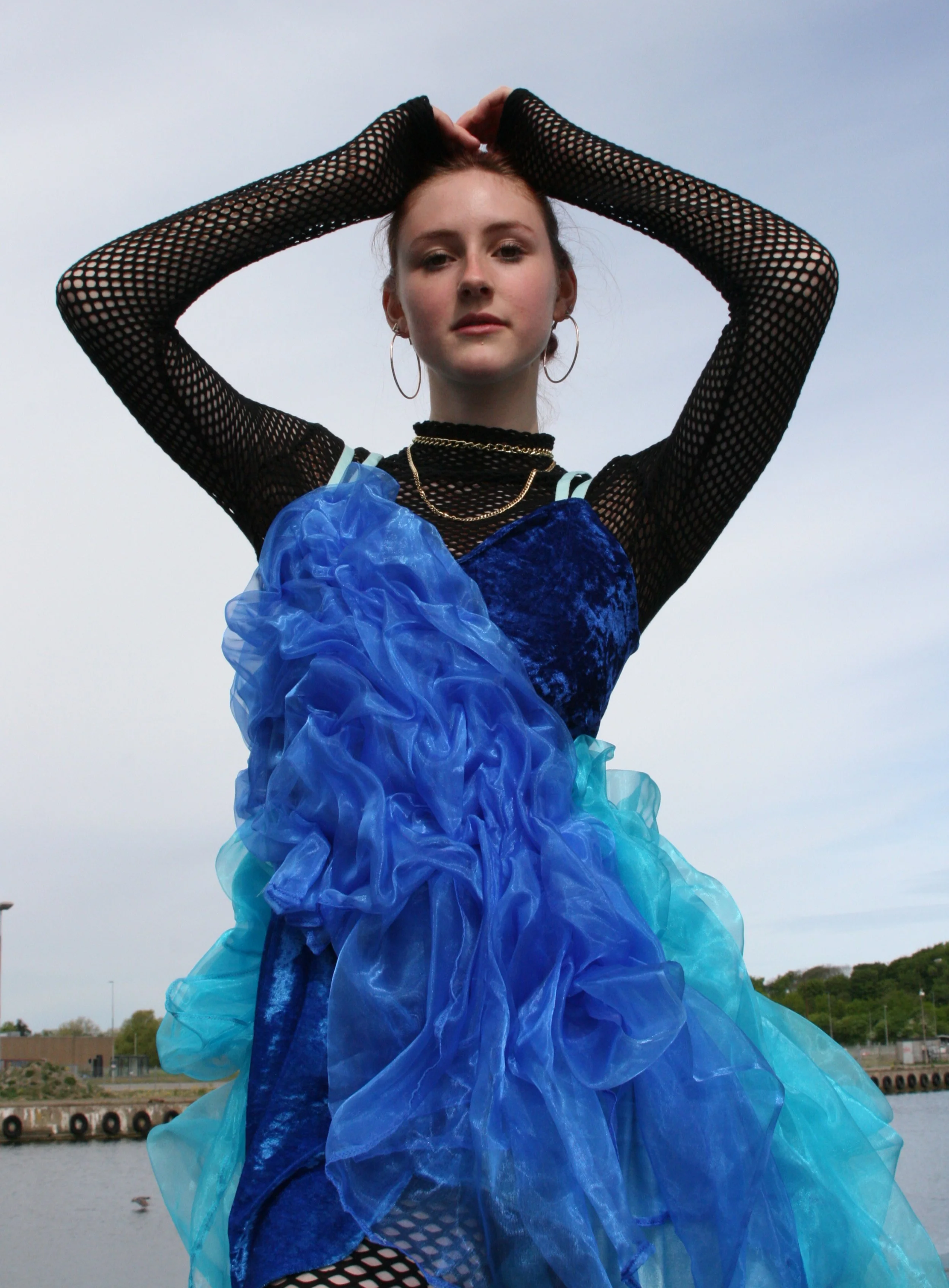 A young woman with auburn hair tied back, wearing a black fishnet long-sleeve top, a velvet blue dress, and layered gold necklaces, standing outdoors by the water with her arms raised above her head, gazing into the camera.