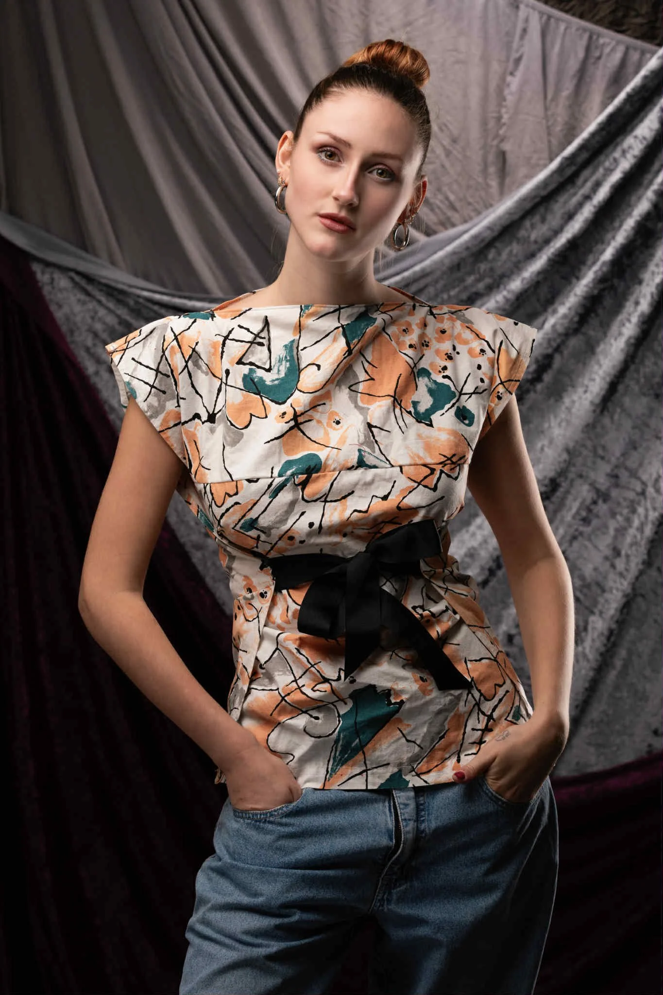 A young woman with red hair in a bun, wearing a colorful patterned top with a black bow and blue jeans, standing against a backdrop of draped fabric.