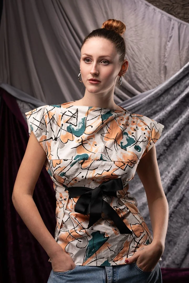 A young woman with red hair tied in a bun, wearing hoop earrings, a colorful patterned top with a black ribbon belt, and blue jeans, standing against a backdrop of textured gray and purple fabric.