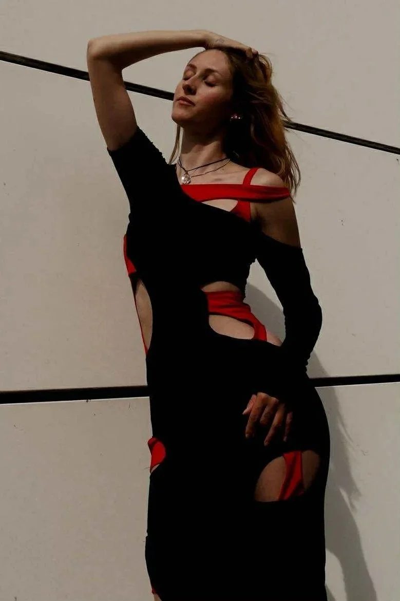 A woman wearing a black and red dress with cutouts, posing with her eyes closed and one hand on her head, standing against a plain white wall with black lines.