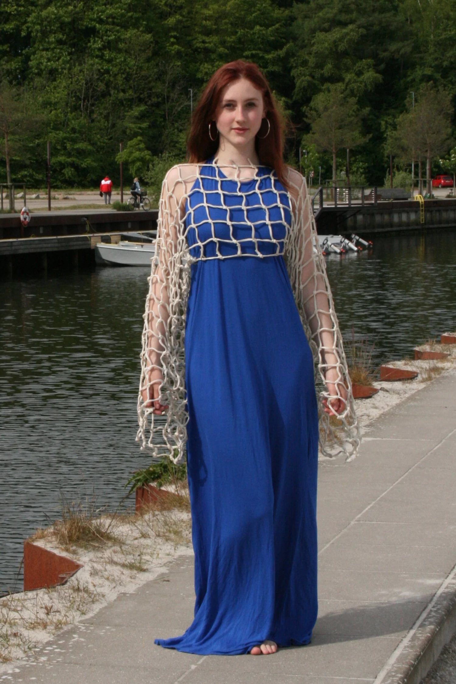 A woman with red hair standing near a body of water, wearing a long blue dress and a beige netted cover-up with large hoop earrings.
