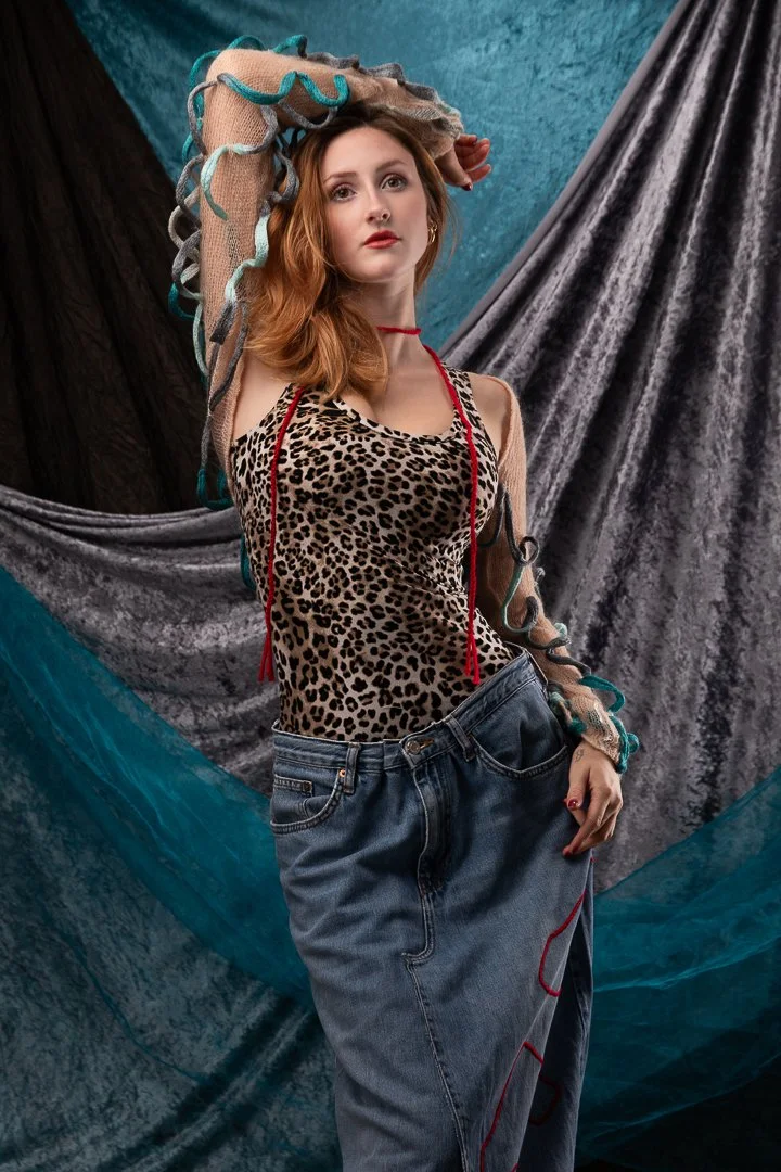 A young woman with red hair wearing a leopard print top and loose denim jeans standing against a textured backdrop with draped fabric.