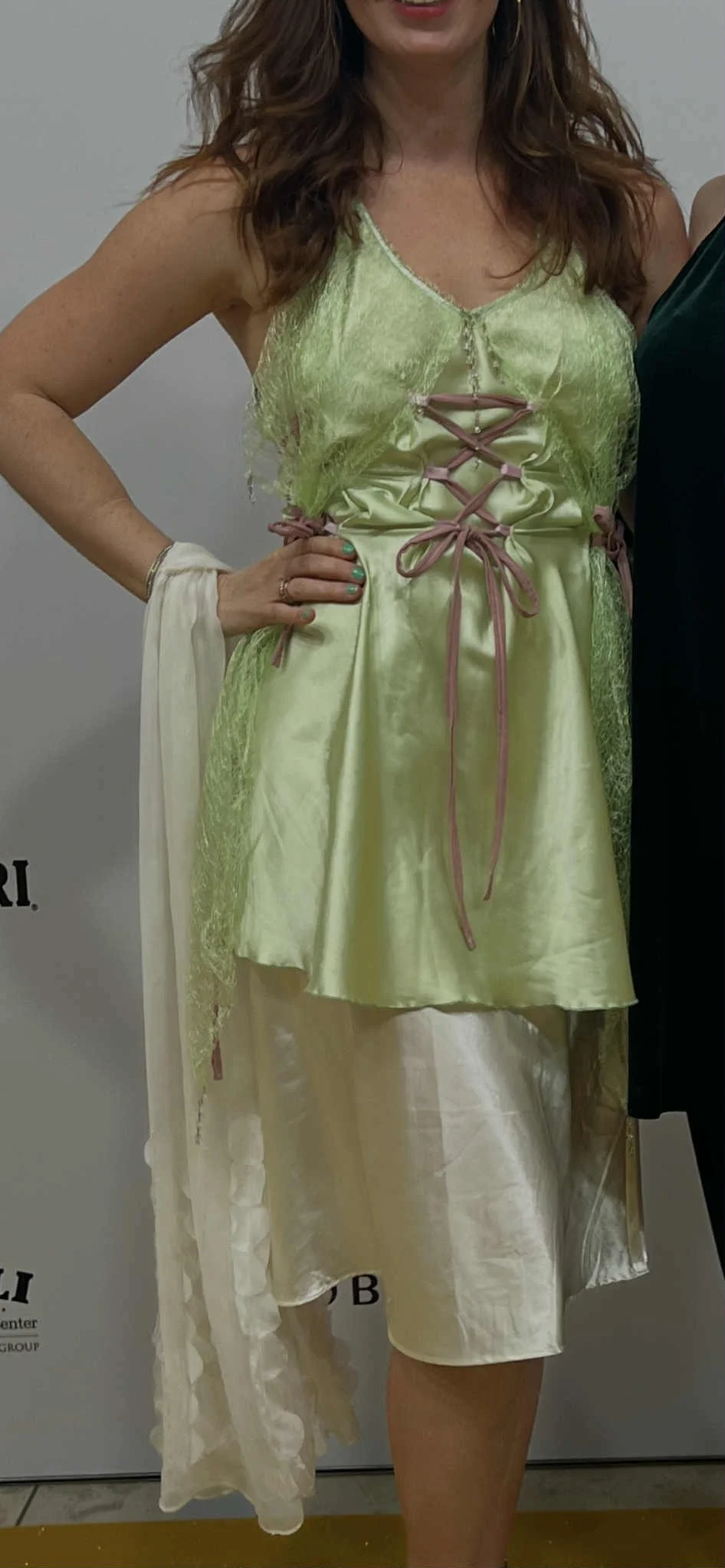 A woman wearing a yellow satin dress with pink lace-up detail in front, layered with a matching green lace overlay, and cream-colored satin skirt underneath.