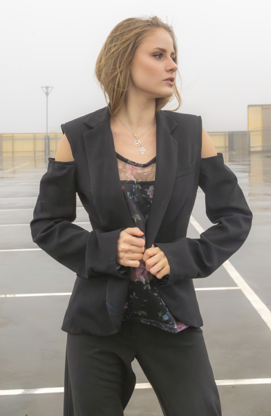 Young woman with blonde hair in a black blazer with cut-out shoulders, standing on a rooftop parking lot with a cloudy sky.