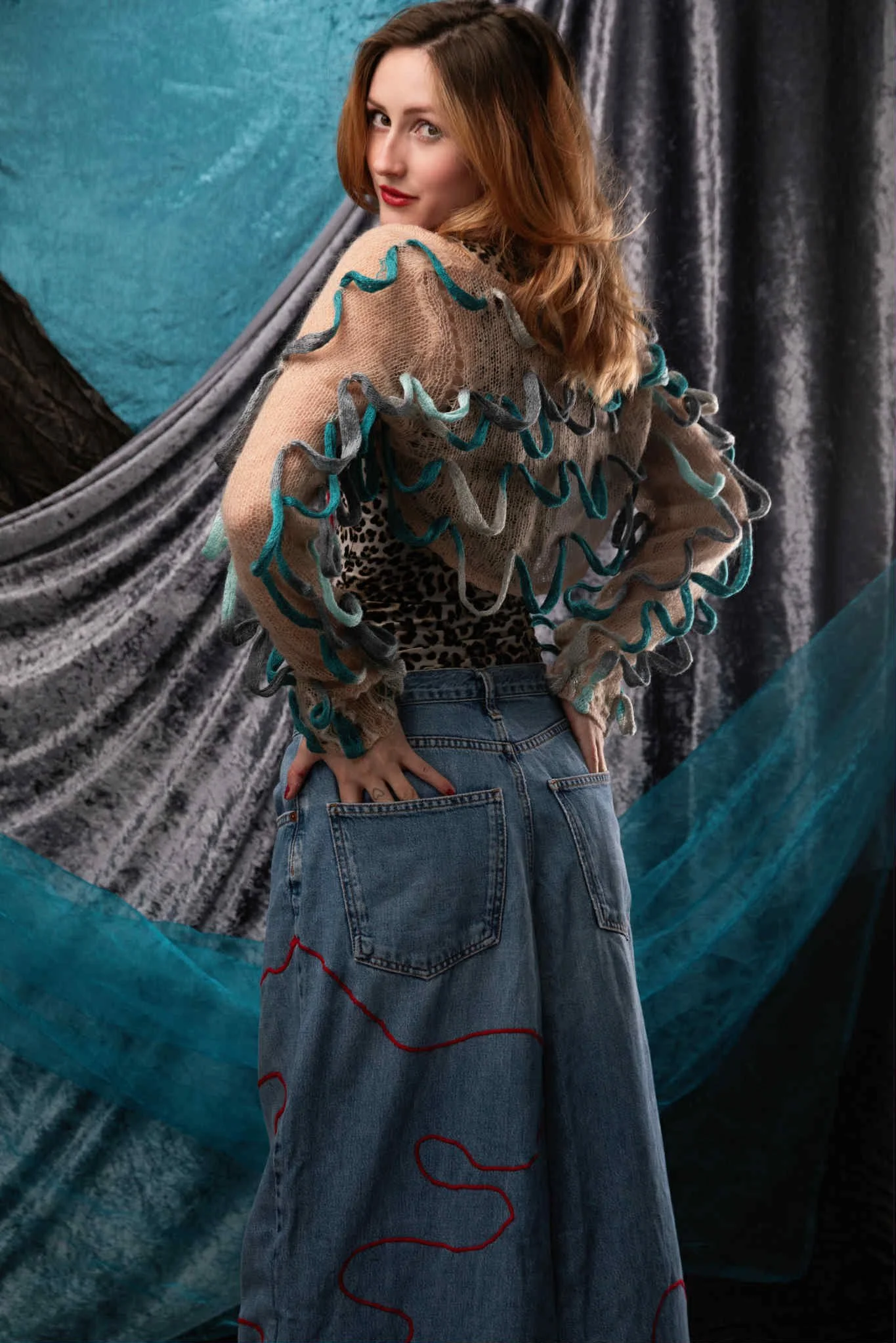 A woman with red hair wearing a beige sweater with ruffled multicolored embroidery, a leopard print top, and wide-leg jeans with red embroidery, standing in front of a textured, layered background of fabric and teal accents.